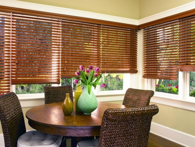 Dining room with wooden blinds, round table, woven chairs, and vase of flowers.