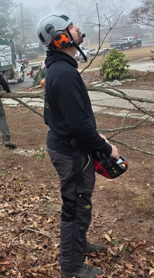 Person in a helmet and dark gear holding a chainsaw in a wooded area, likely after storm cleanup