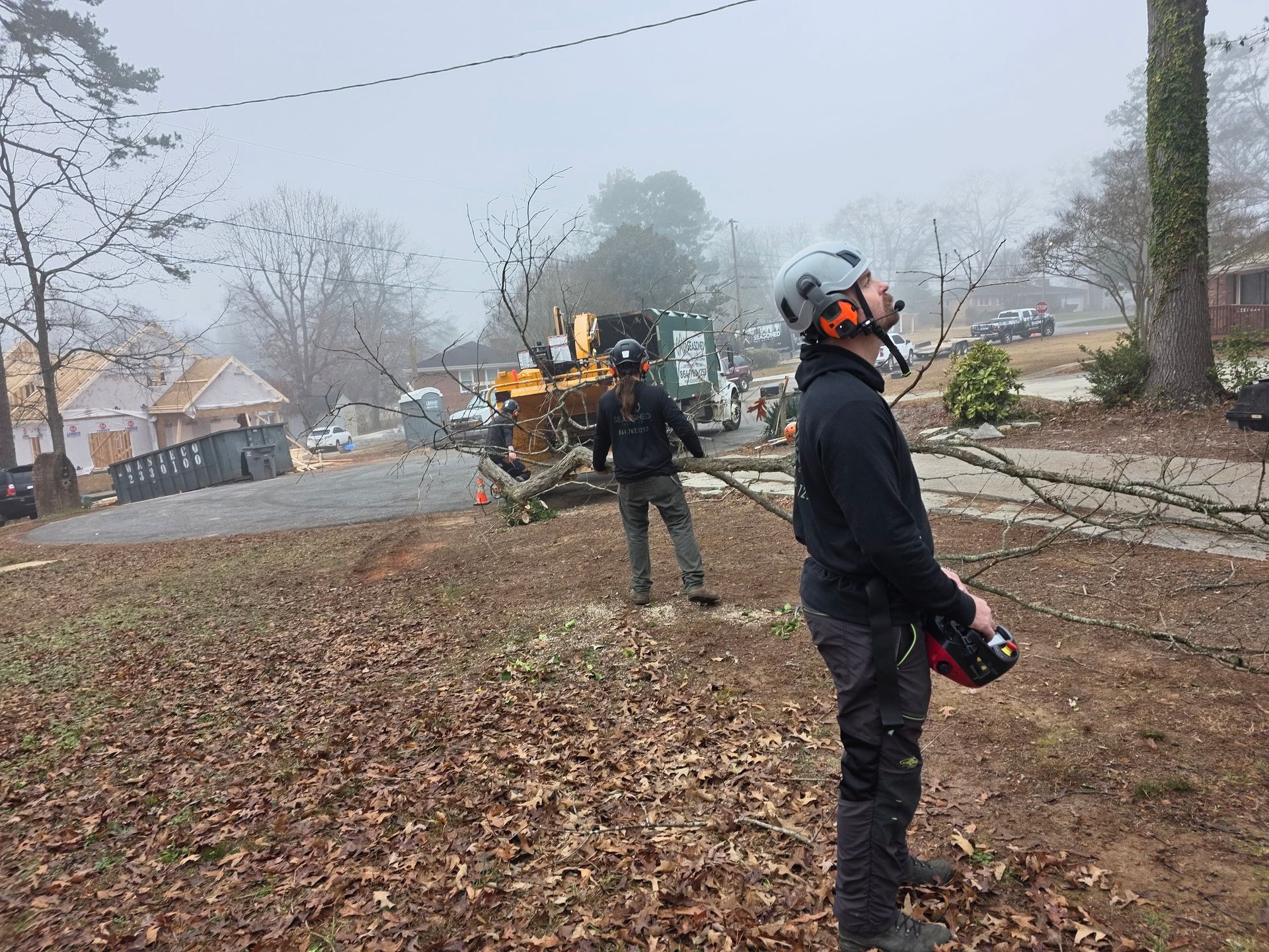 Workers clearing storm debris on a leaf-covered street with a chainsaw and utility truck in foggy weather