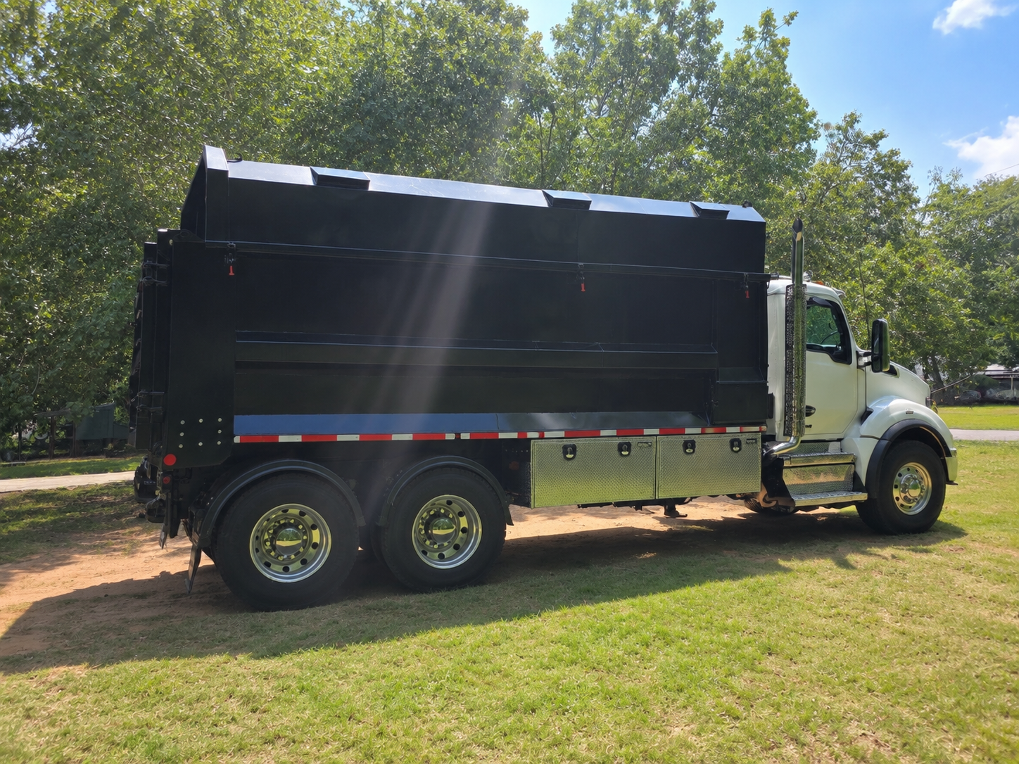 Large black dump truck parked on grass under trees on a sunny day