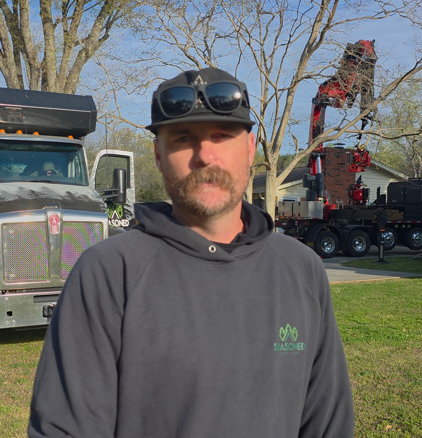 Man in dark hoodie and cap standing outdoors near a truck and trees on a sunny day
