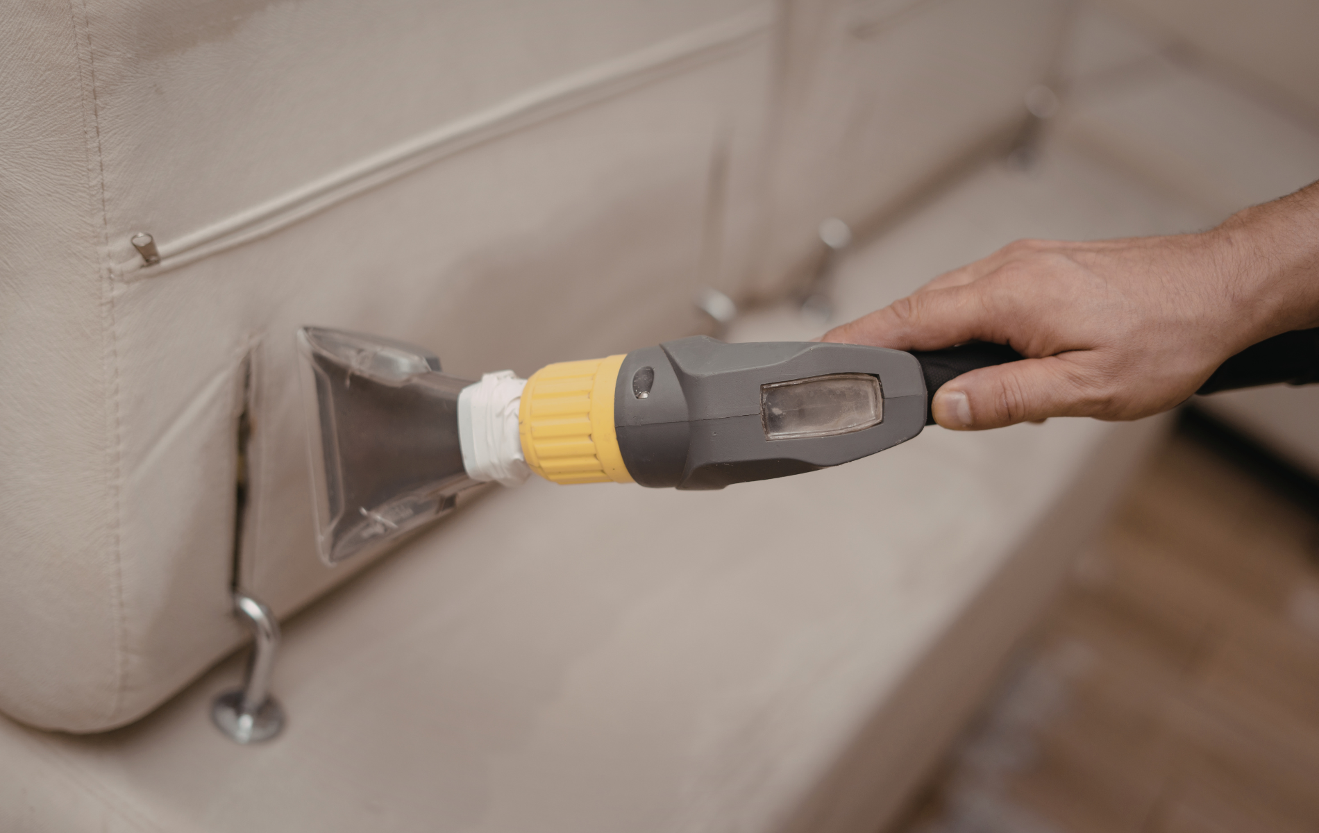 A person uses a handheld upholstery cleaning tool to deep clean a light-colored fabric sofa.