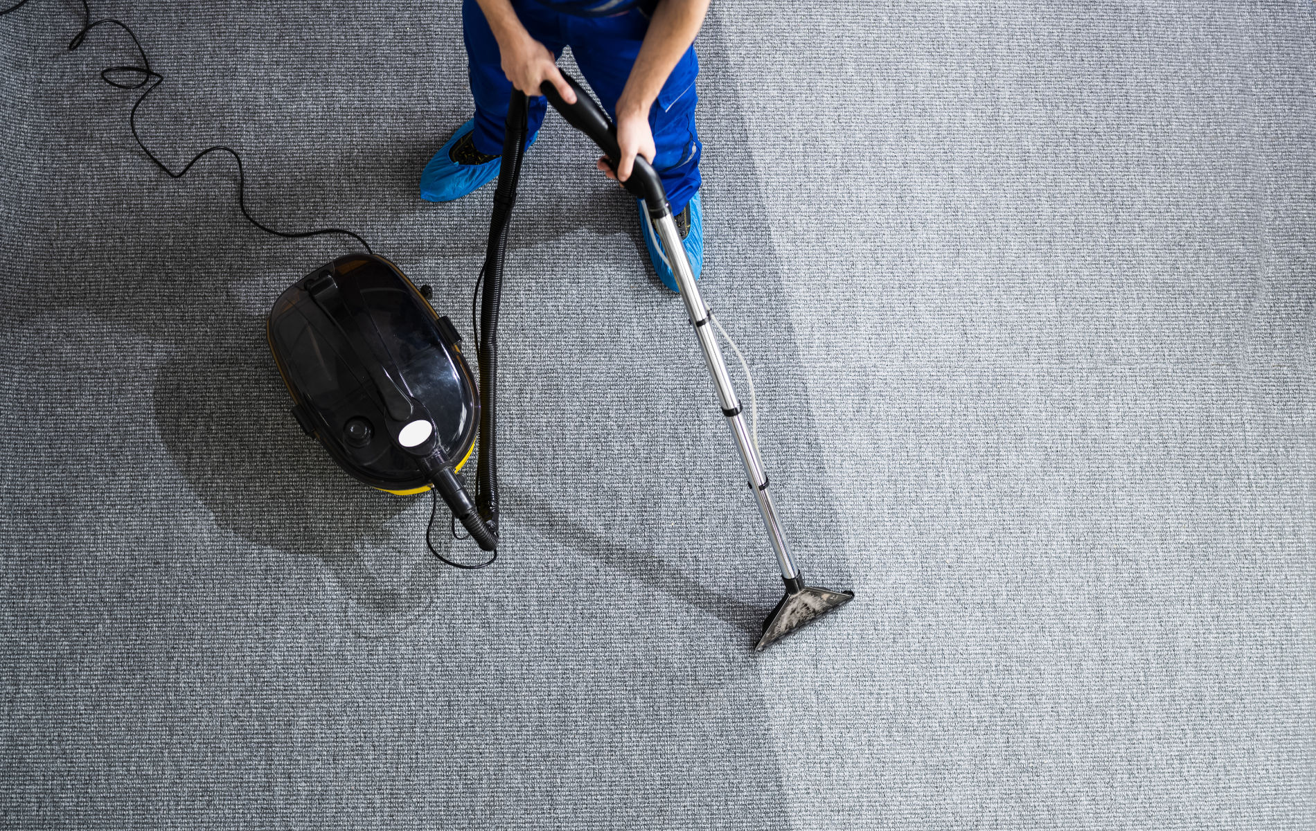 A person in blue pants cleans a textured gray carpet using a black canister vacuum.