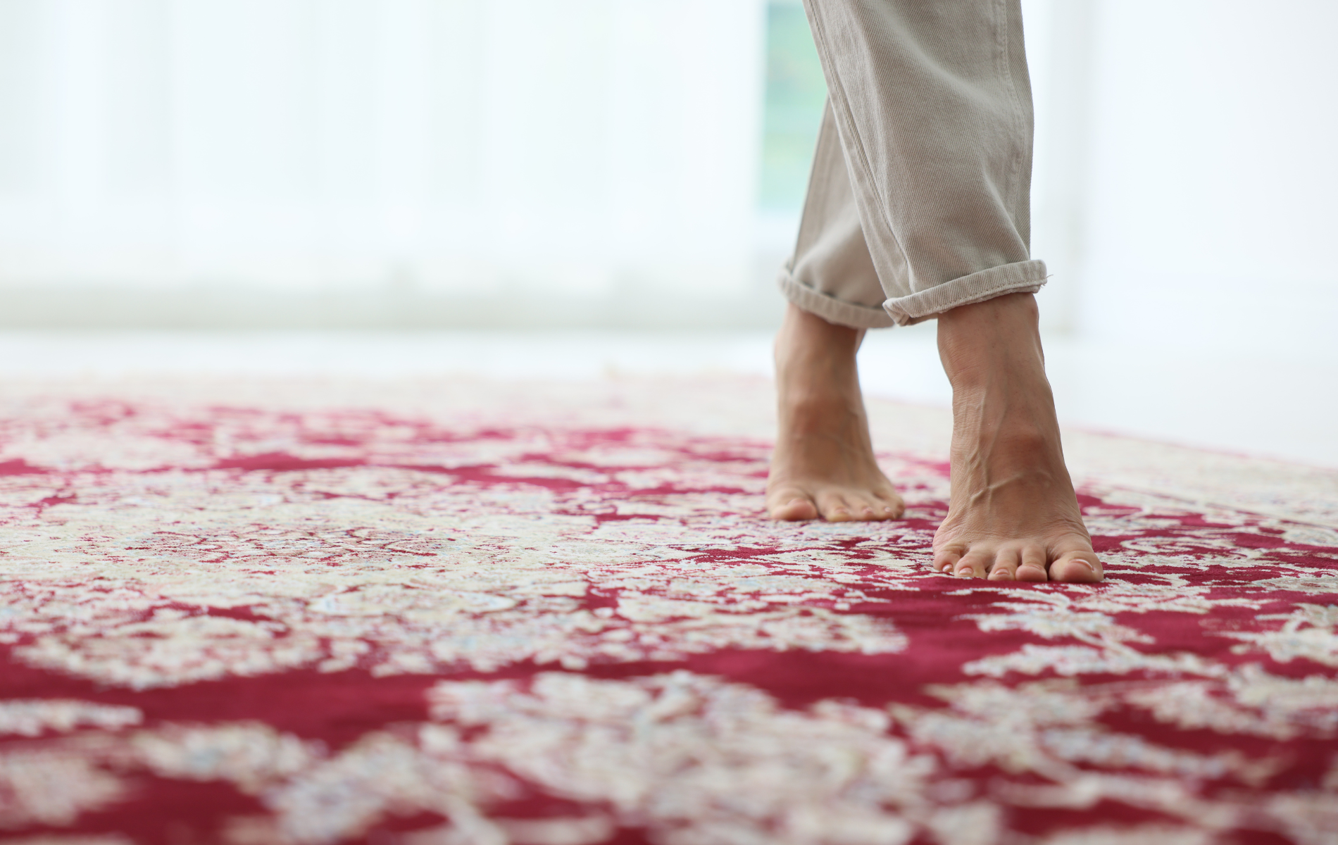 Bare feet walking on a patterned red and white rug, with the hem of light-colored trousers visible above.