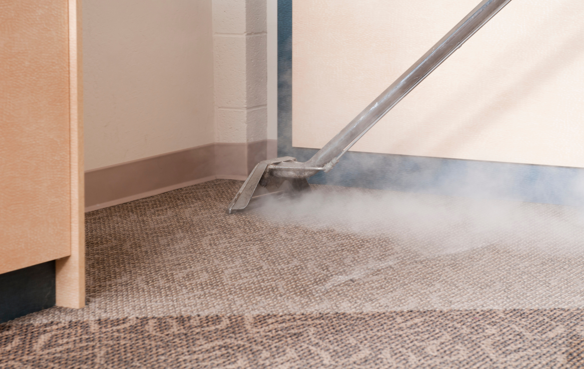 A carpet cleaning wand releasing steam onto a patterned gray carpet near a beige wall.