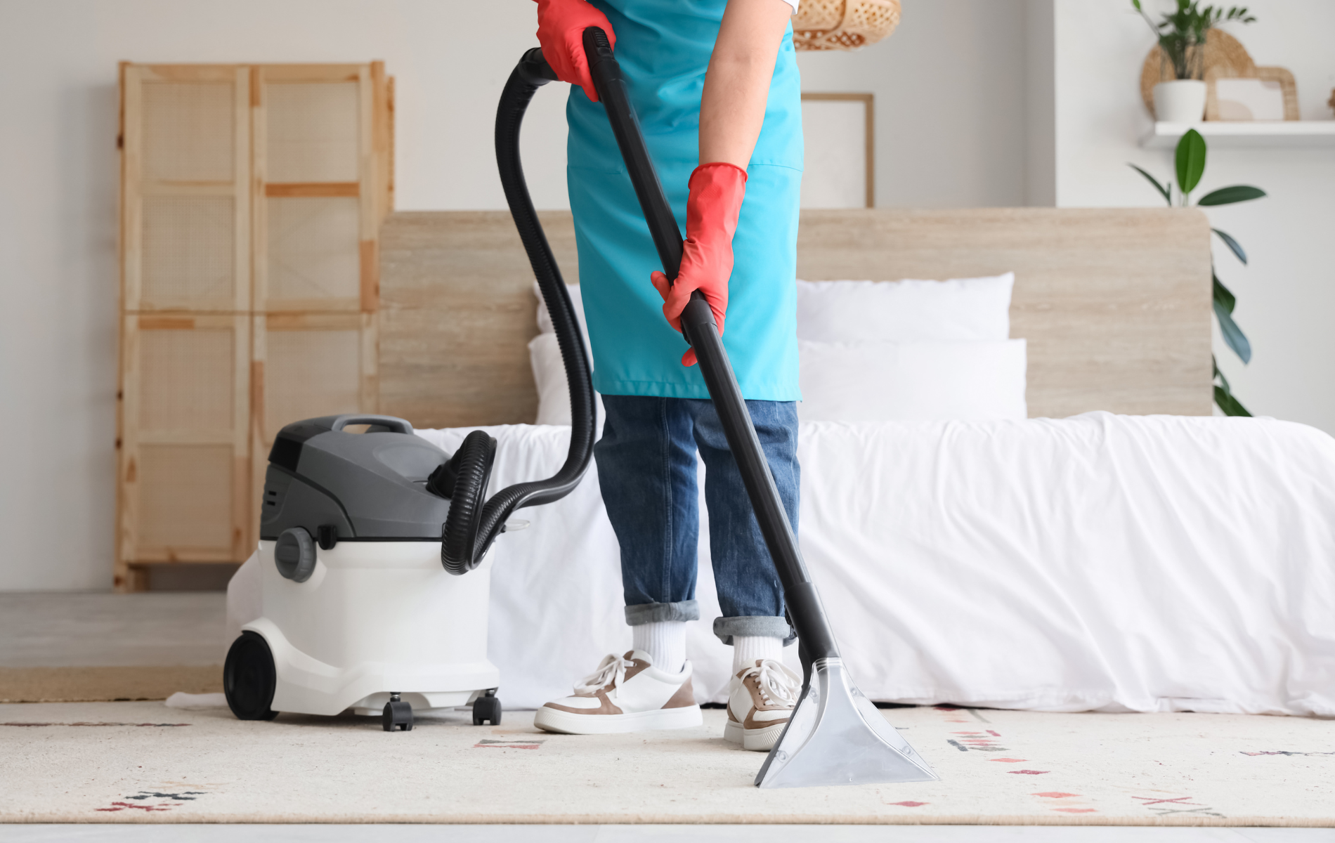 A person in a blue apron and red gloves cleans a white carpet with a vacuum machine in a bright bedroom.