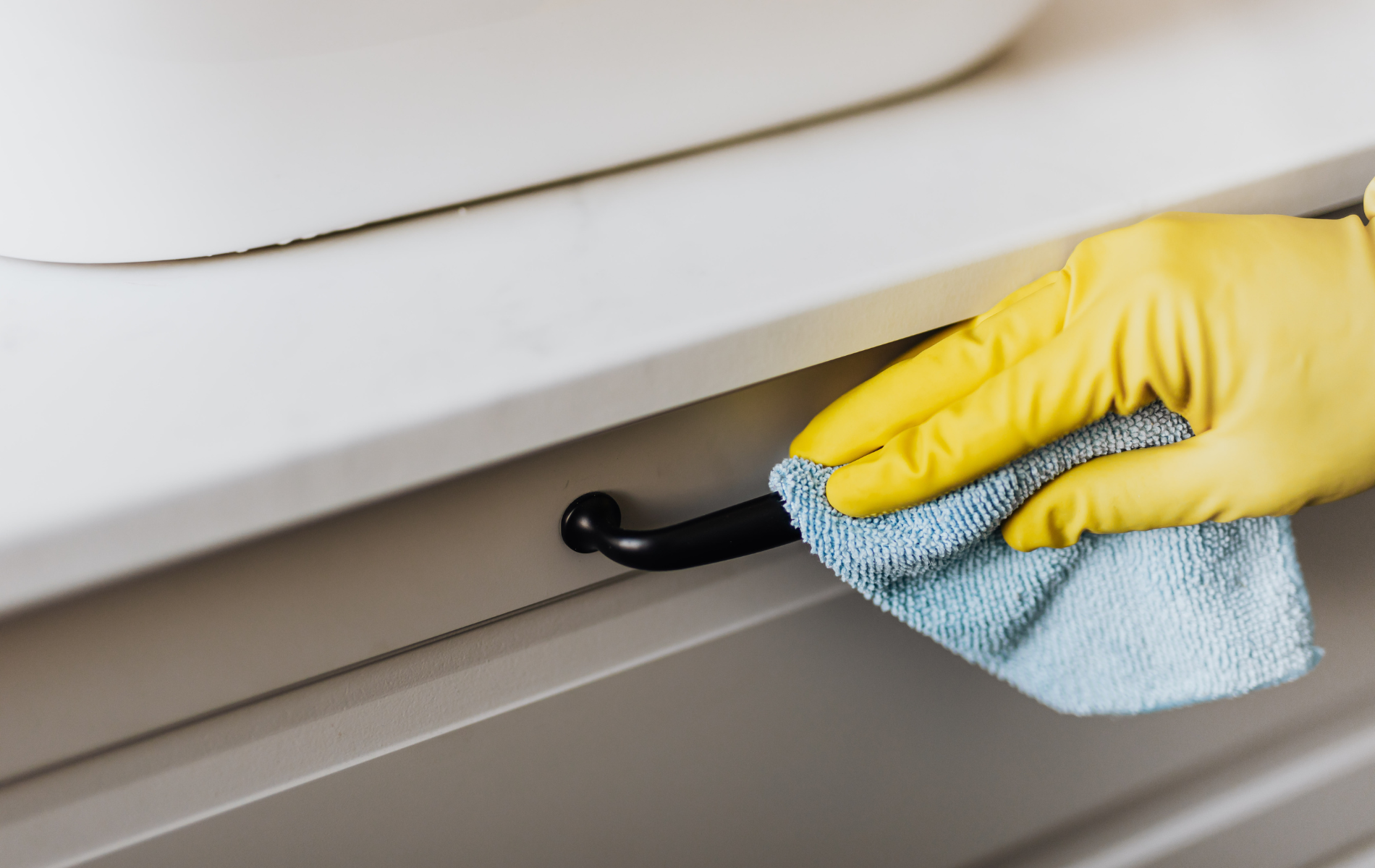 A gloved hand wearing a yellow rubber glove cleans a dark cabinet handle with a light blue cloth.