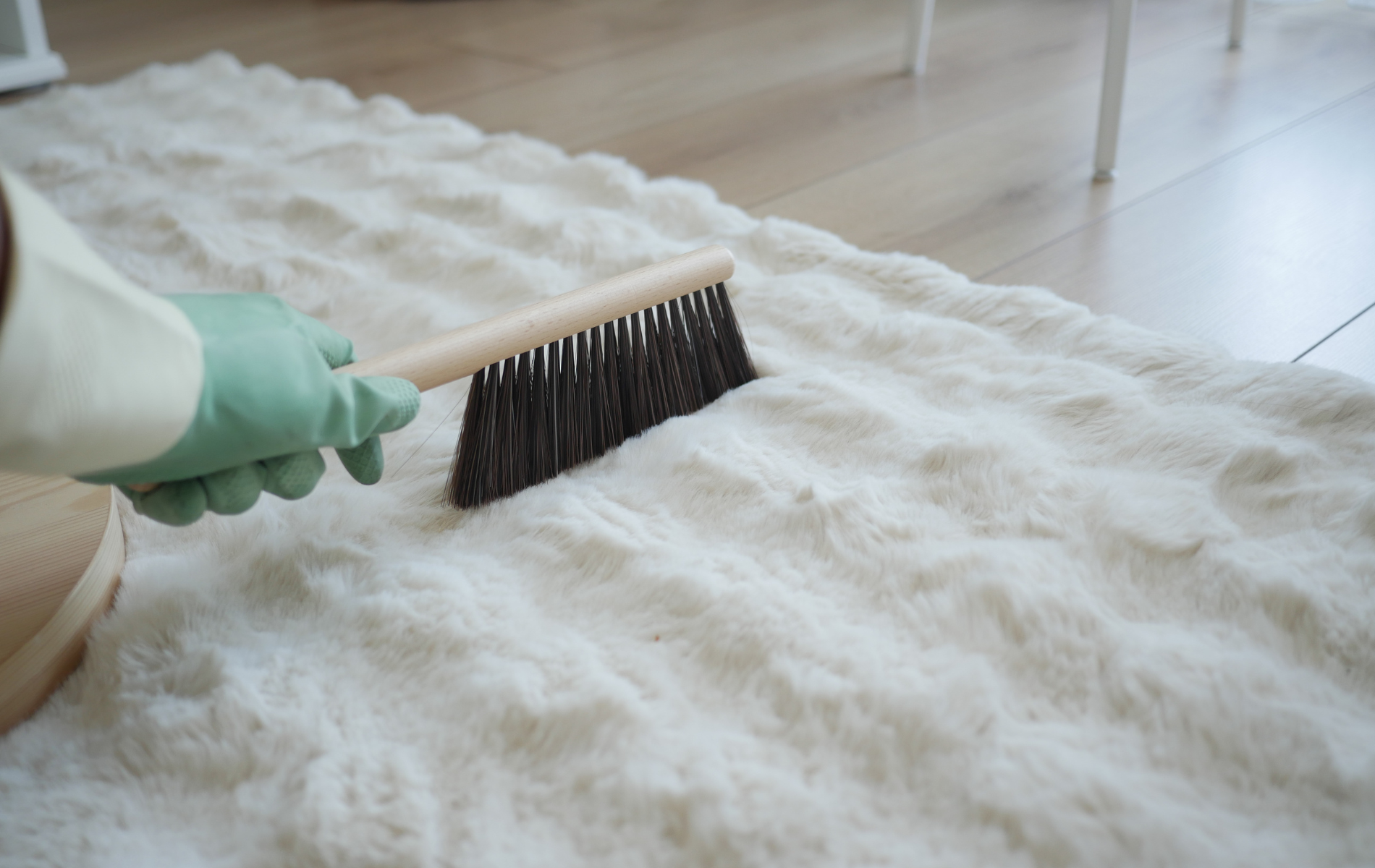 A hand wearing a green glove uses a stiff-bristled brush to groom the surface of a fluffy, white faux fur rug.