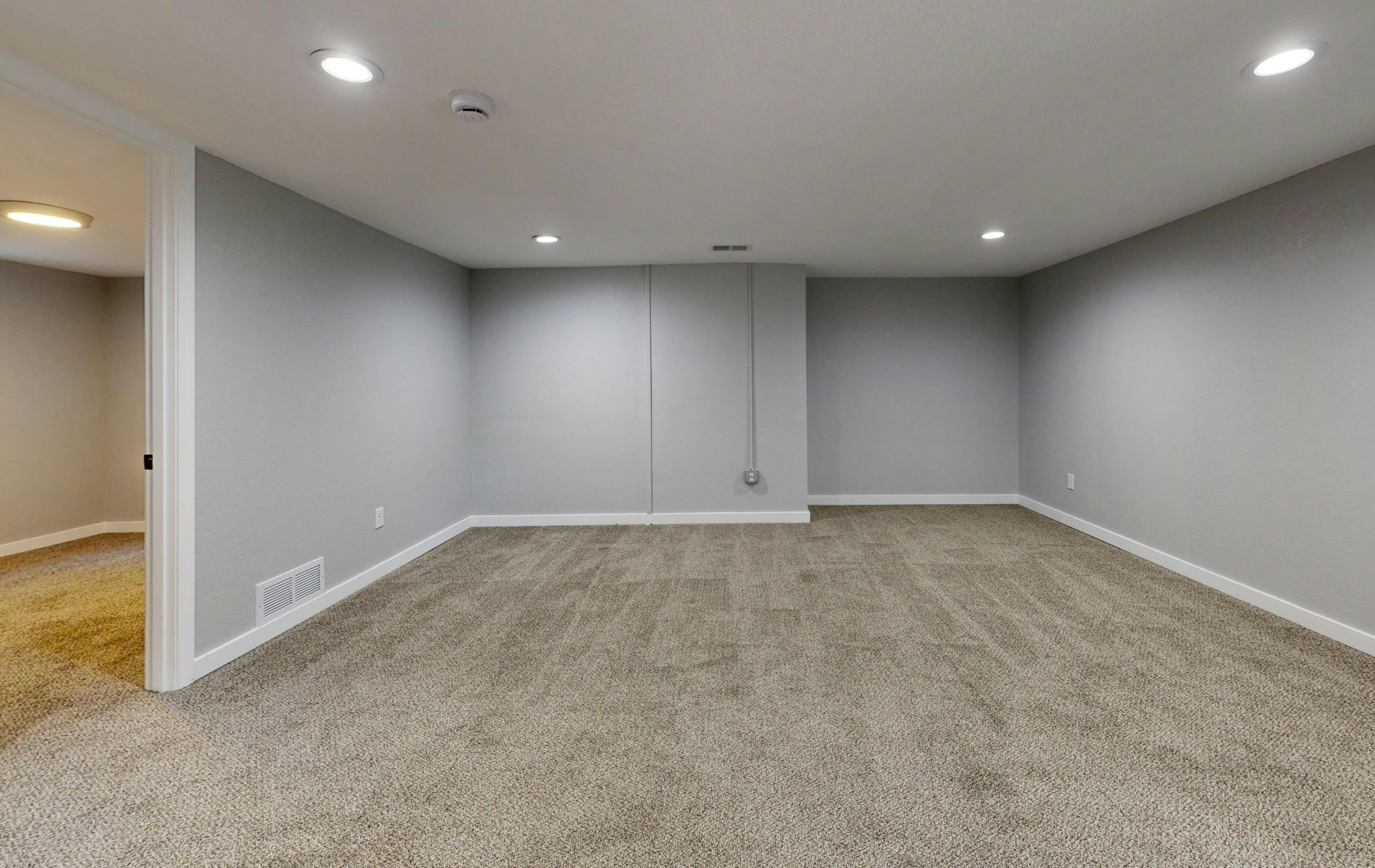 An empty basement room with grey walls, white trim, beige carpeting, and recessed ceiling lighting.