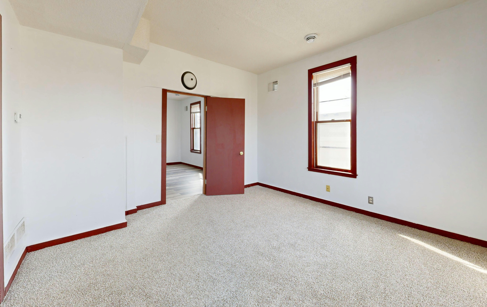 An empty room with white walls, carpeted floors, a window with a red frame, and an open doorway to another room.
