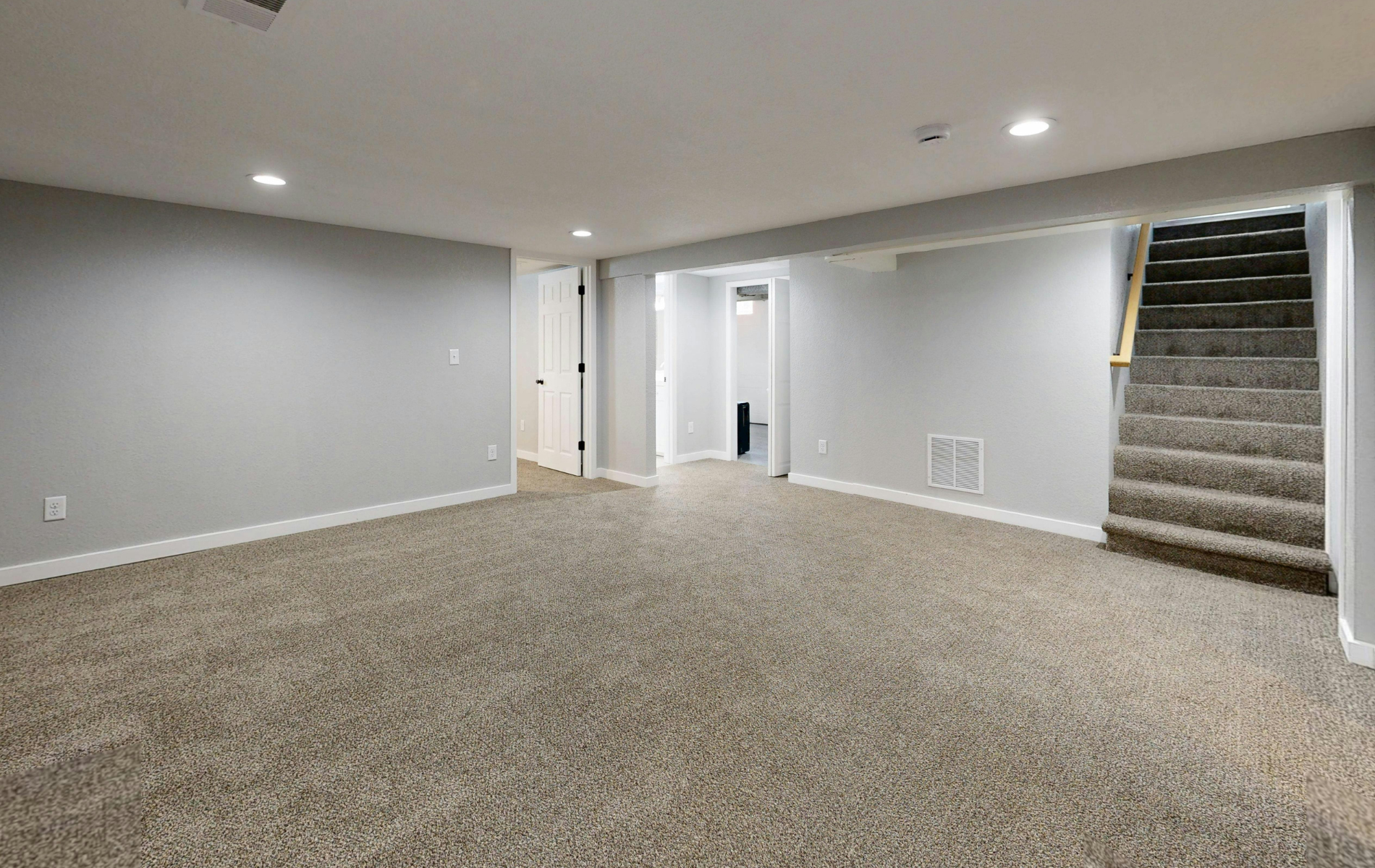 A modern, empty basement with light gray walls, neutral wall-to-wall carpeting, recessed ceiling lighting, and stairs.