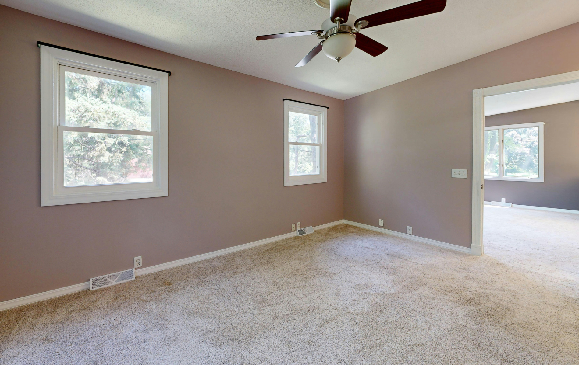 An empty room with light tan walls, carpet flooring, a ceiling fan, and two windows leading to an adjacent room.