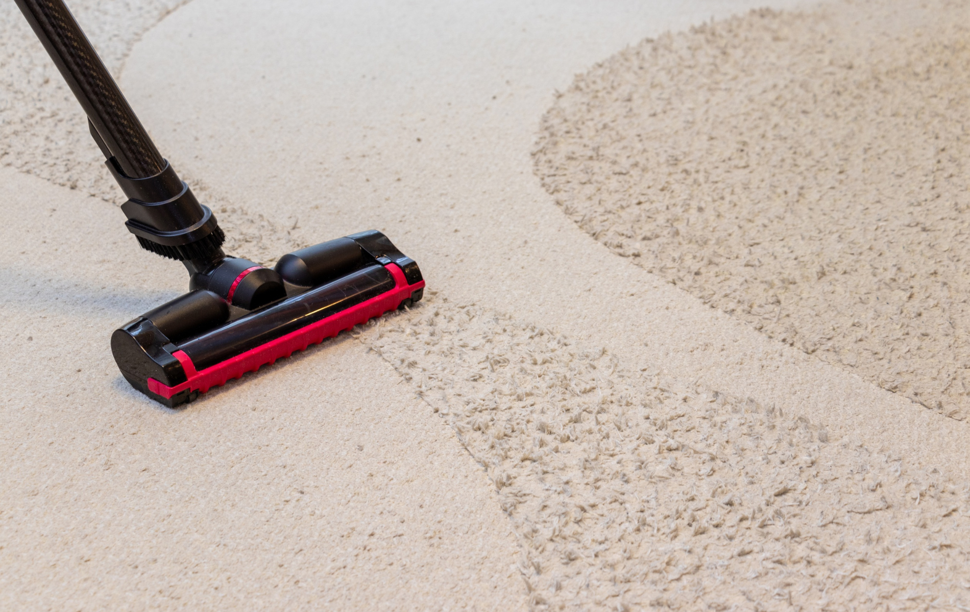 A black and red vacuum cleaner head glides across a beige carpet, leaving behind a clean, smooth trail.