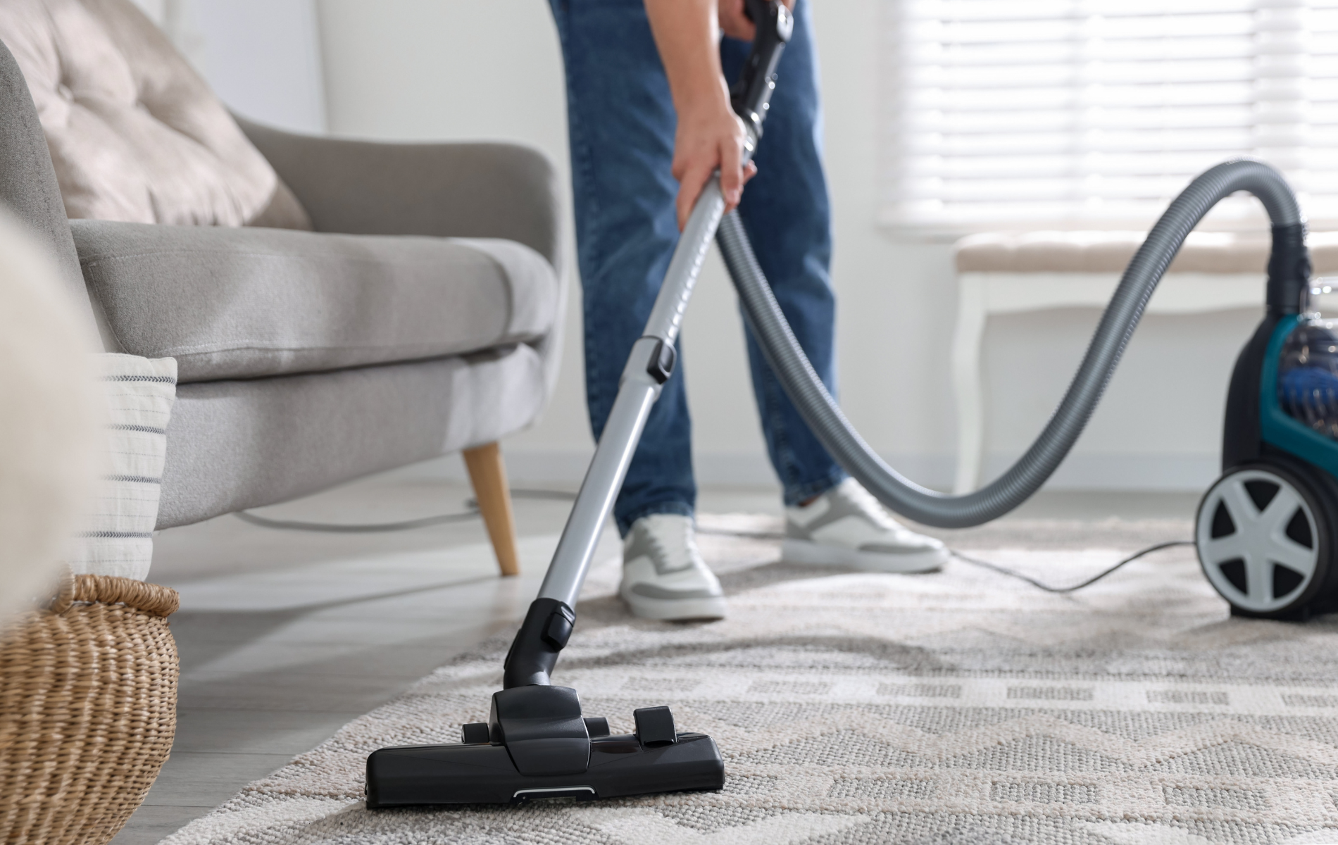 A person vacuums a patterned area rug in a brightly lit living room with a grey sofa.