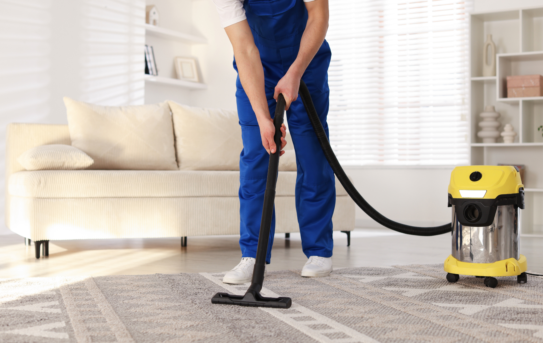A person in blue coveralls uses a yellow industrial vacuum cleaner to clean a patterned rug in a bright, modern living room.