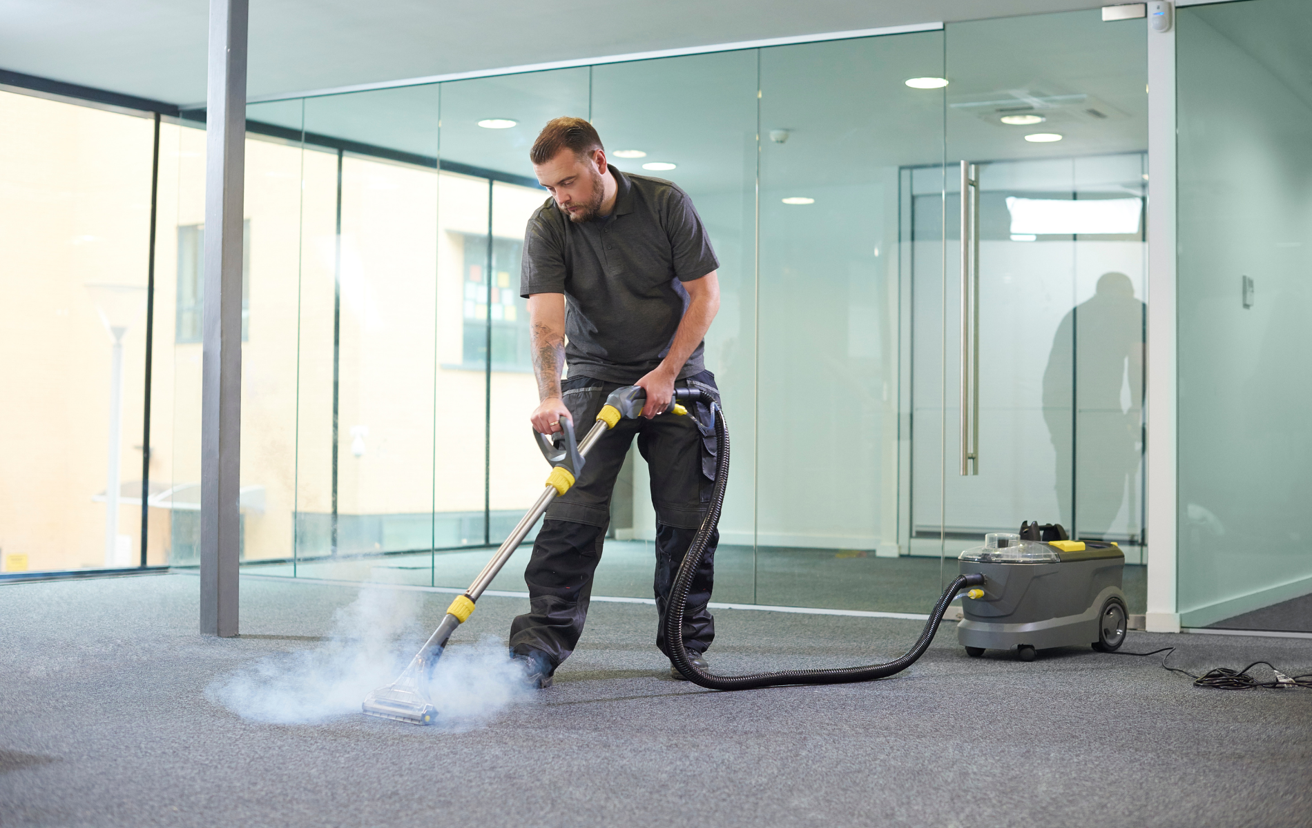 A person in work clothes uses a steam cleaner on a gray office carpet in a modern, glass-walled building.