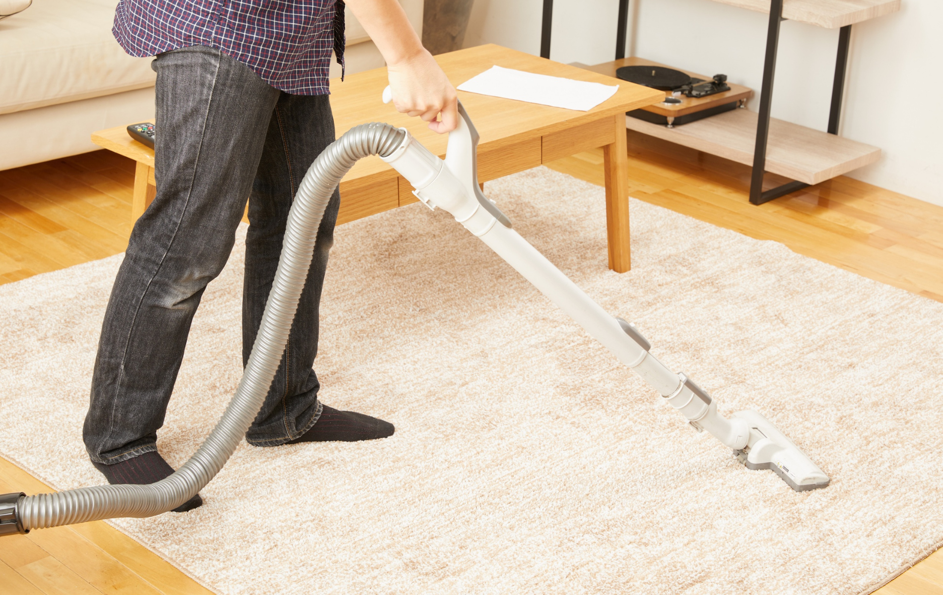 A person vacuuming a light-colored rug in a living room near a wooden coffee table and shelves.