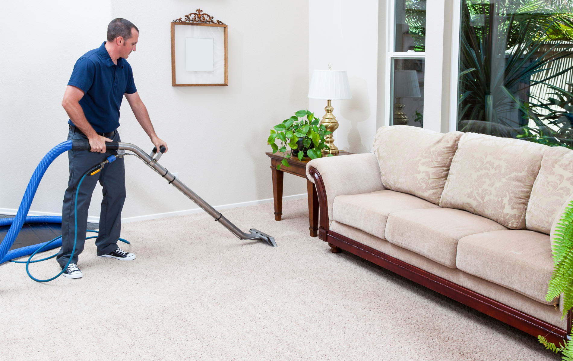 A person in a blue shirt cleans a carpeted living room floor with an industrial vacuum wand near a beige sofa.