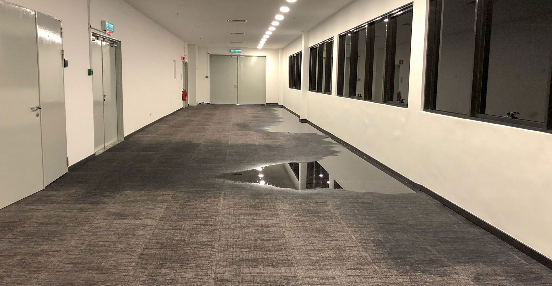 A wide, empty hallway with gray carpeting and a large, reflective puddle of water pooled on the floor near the windows.