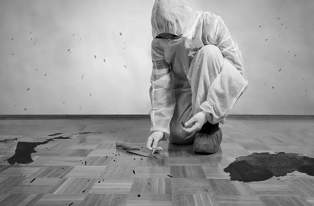 A person in a protective white coverall suit kneels on a wooden floor, examining dark spills and stains.