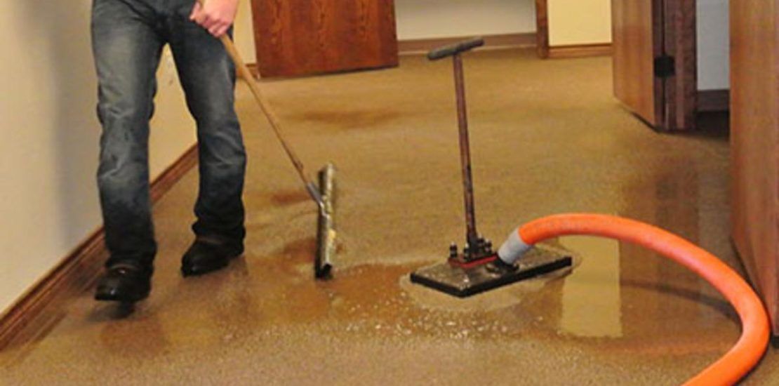 A person uses a floor squeegee and a vacuum extraction tool to remove standing water from a carpeted hallway.
