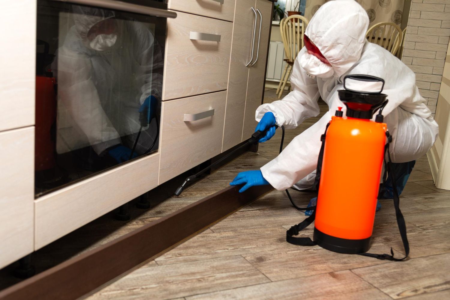 Person in protective suit spraying under a kitchen cabinet with an orange tank.