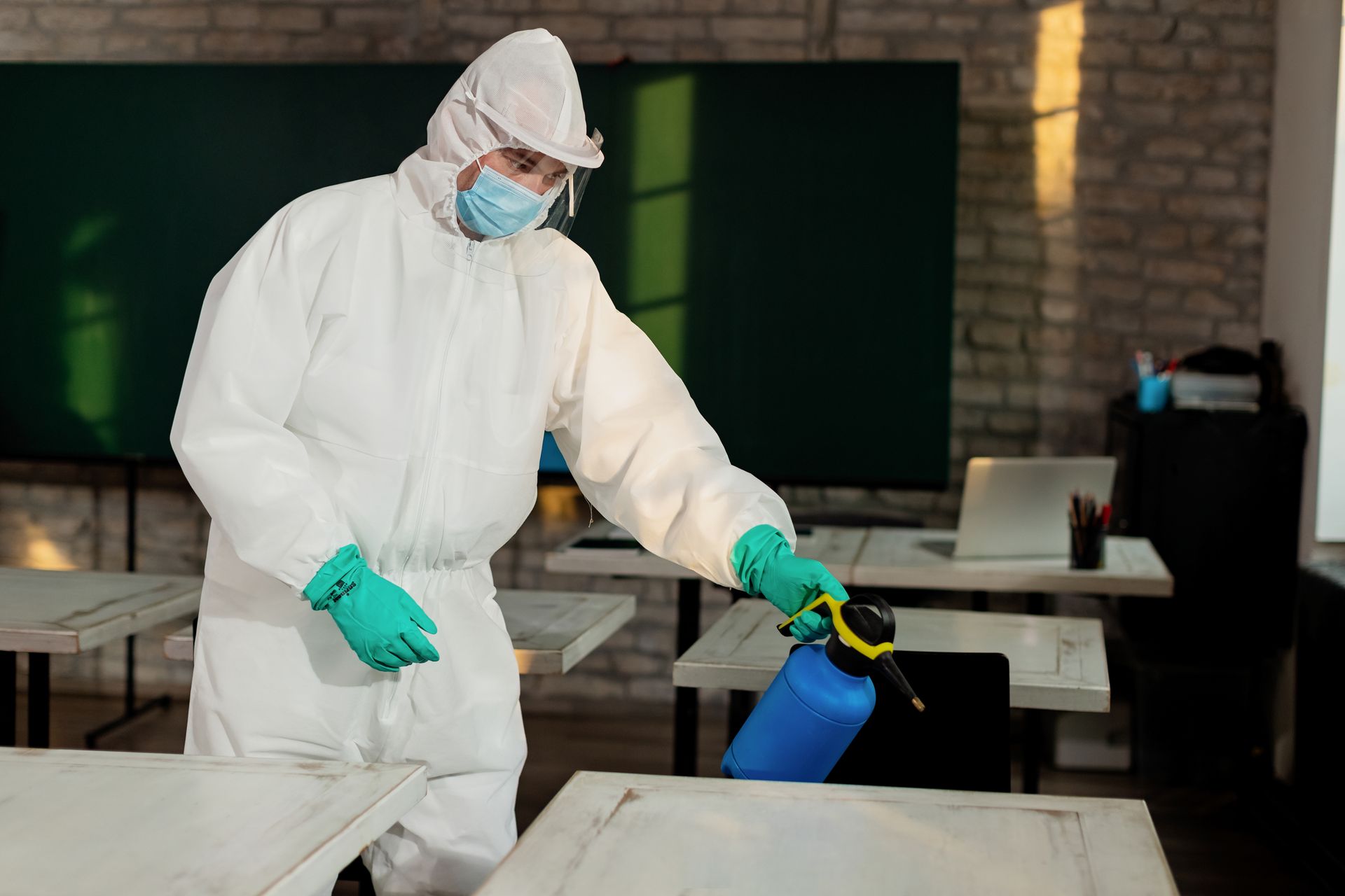 Person in protective suit sanitizing a classroom, spraying desks with a blue bottle.