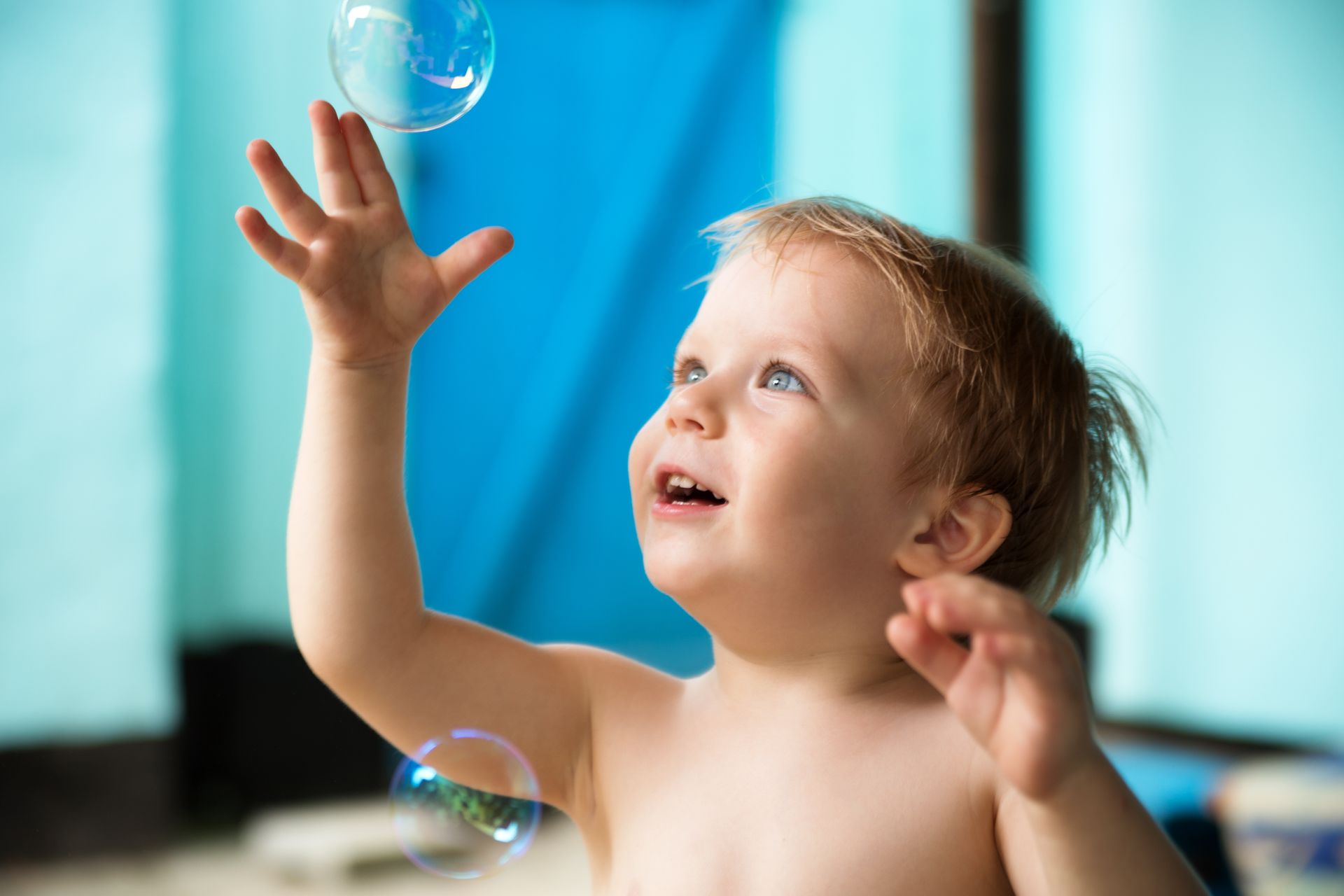 Blond toddler reaches up with glee to catch floating bubbles, set against a blue wall.