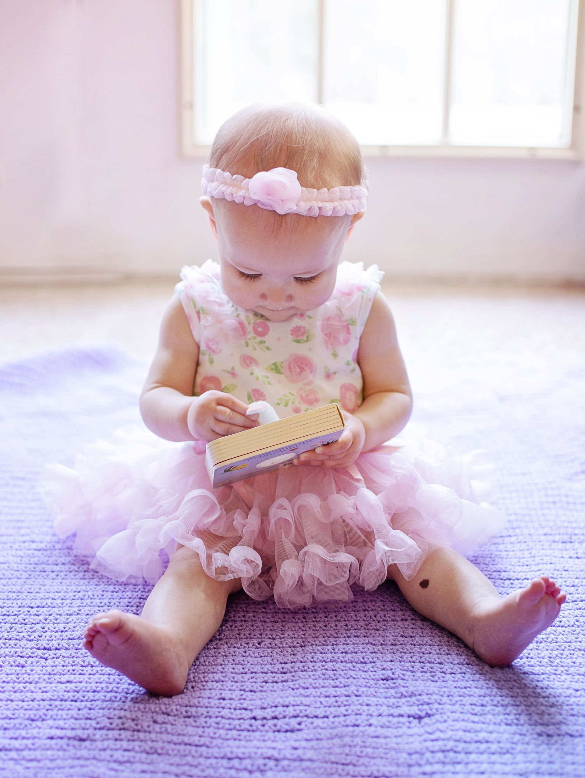 Baby in pink dress and headband playing with a small box, sitting on a purple rug.