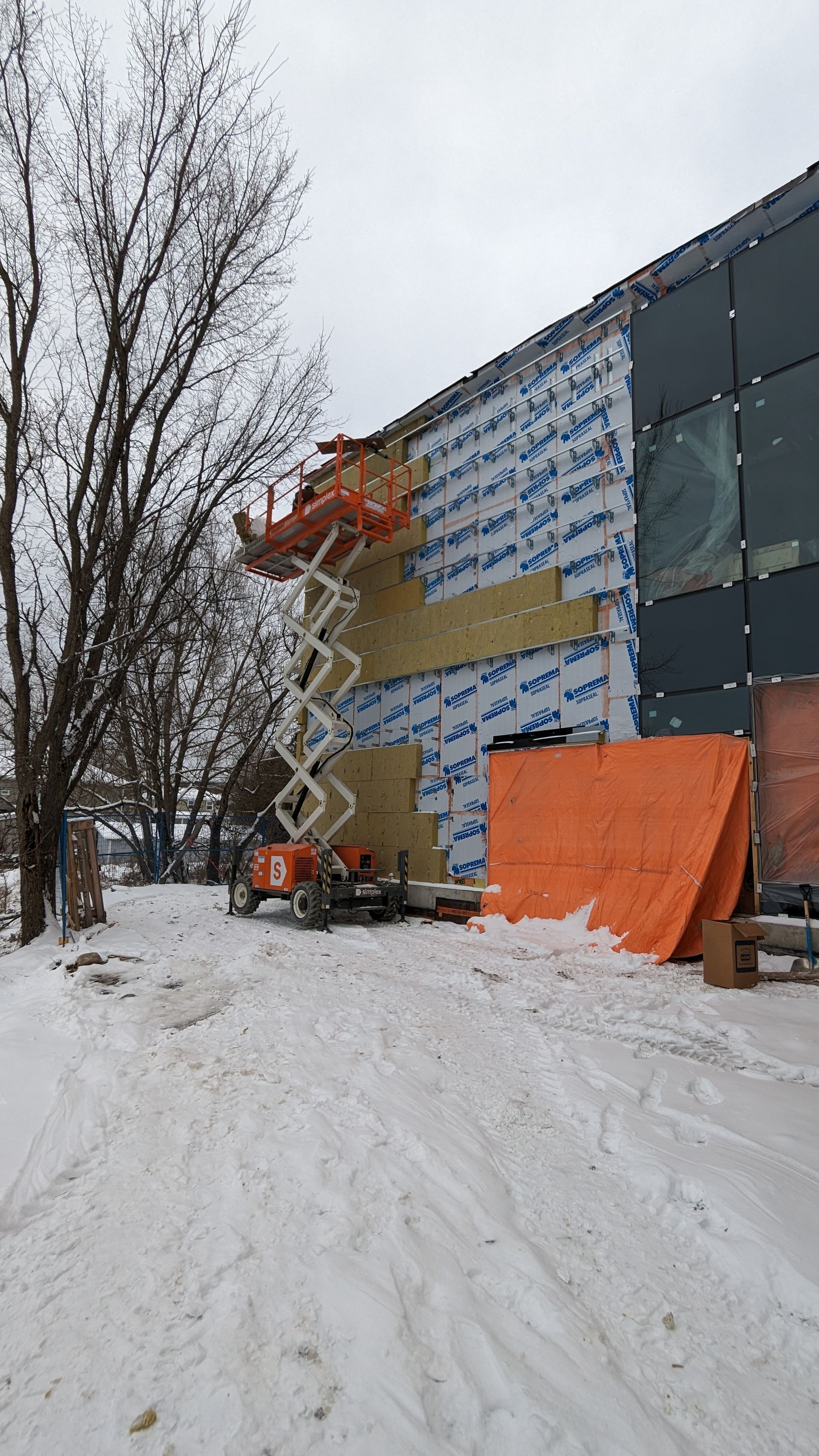 Un ouvrier du bâtiment, sur une nacelle élévatrice, installe de l'isolant sur la façade d'un bâtiment ; il y a de la neige au sol.