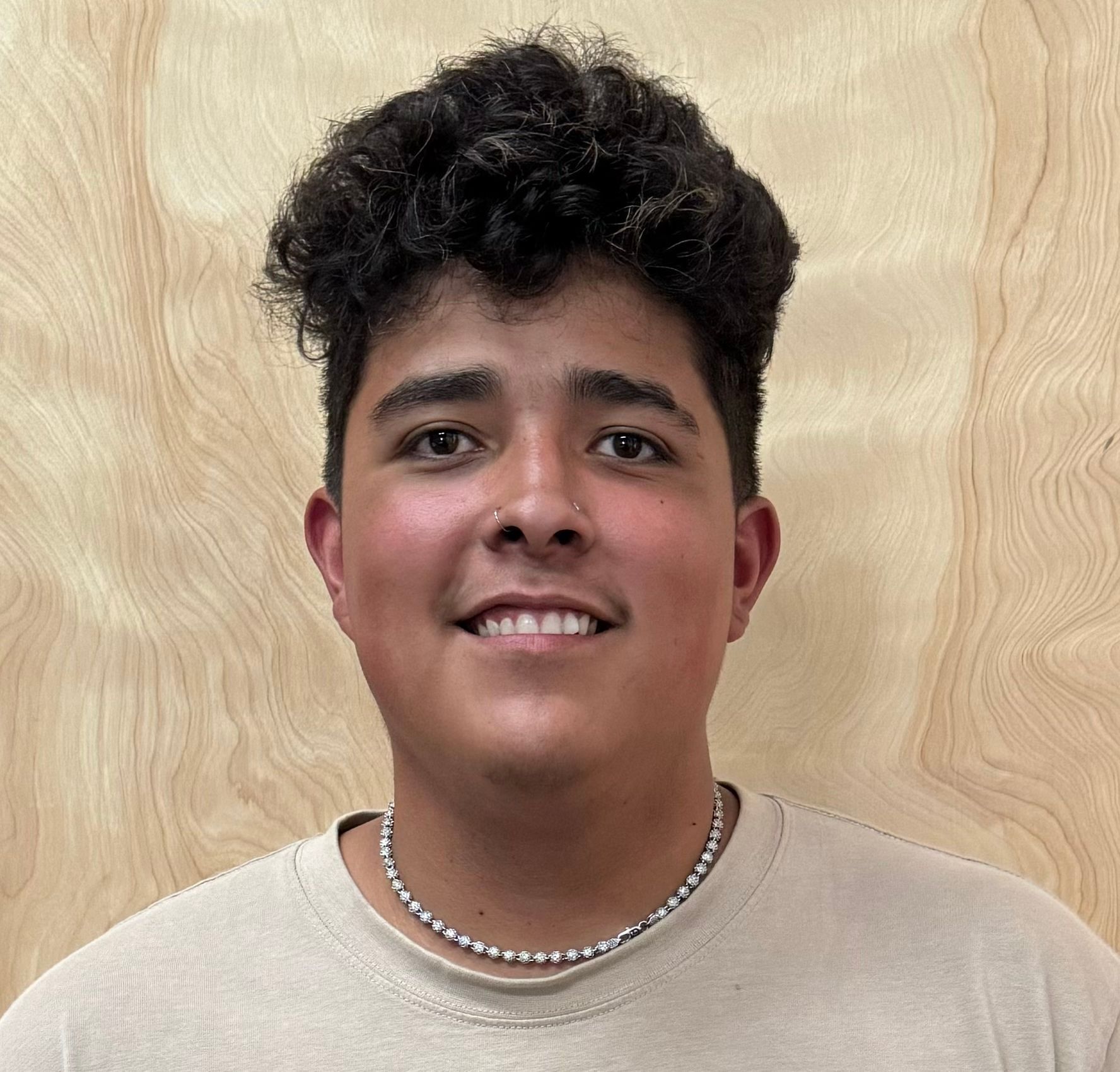 A young man with curly hair is wearing a necklace and smiling.