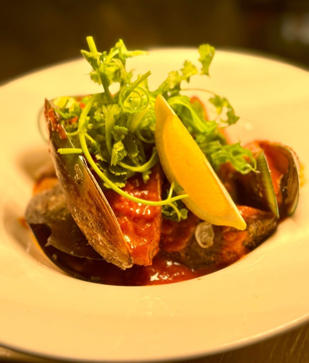 Mussels in red sauce, topped with greens and lemon wedge on a white plate.