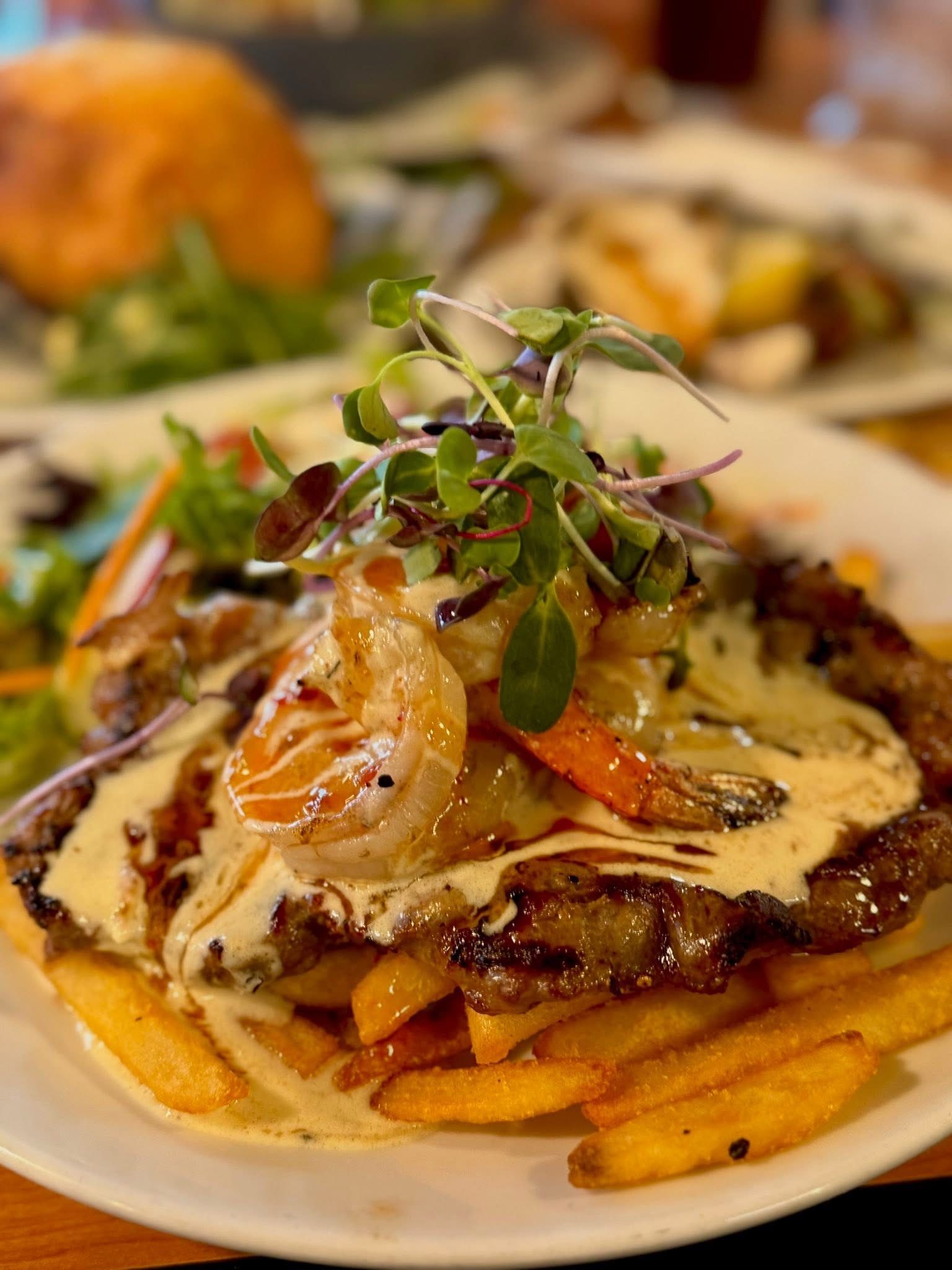 Steak dish with shrimp, fries, and sauce. Garnished with sprouts, served on a white plate.