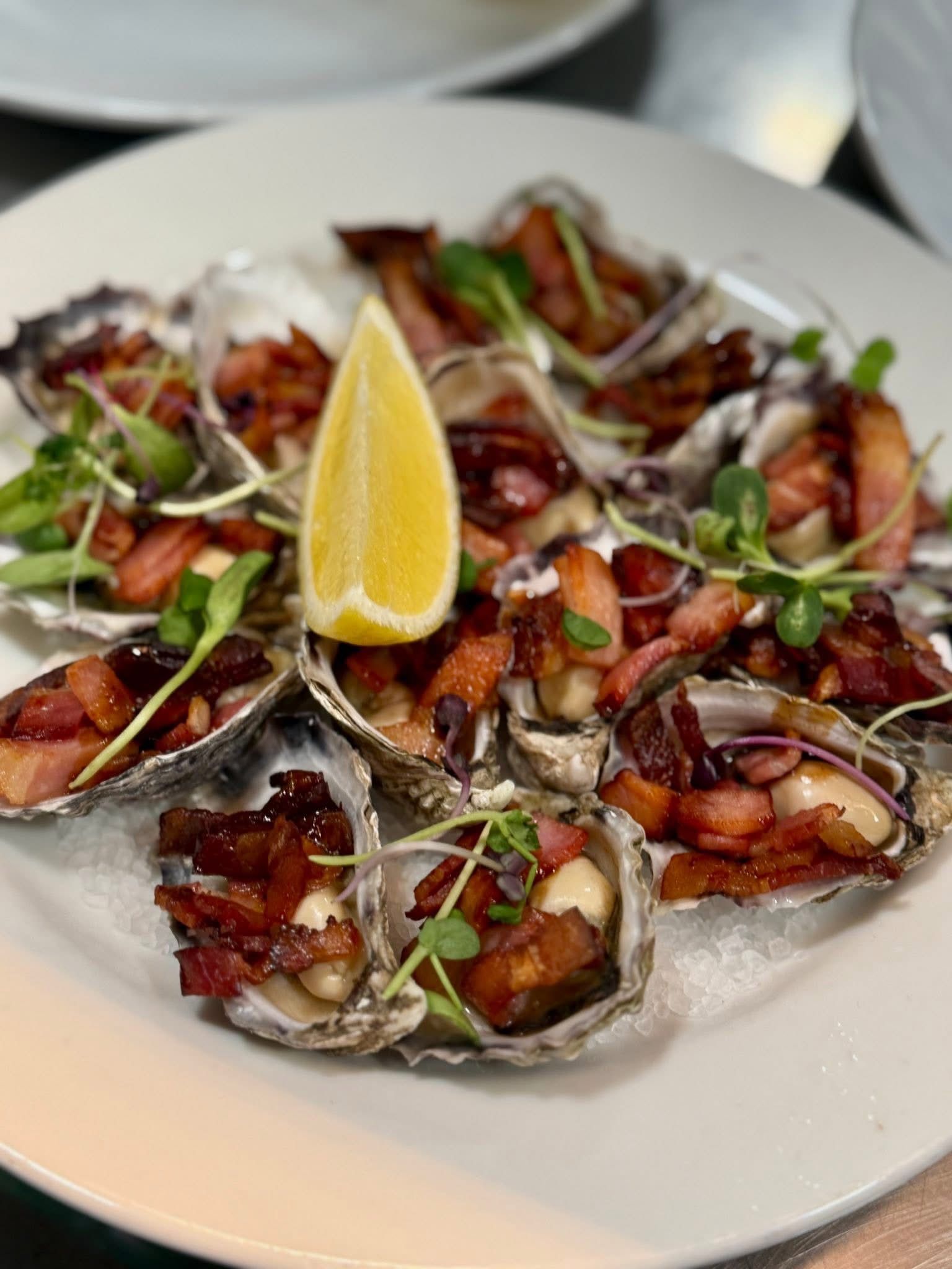 Oysters with bacon on a white plate, garnished with sprouts and a lemon wedge.