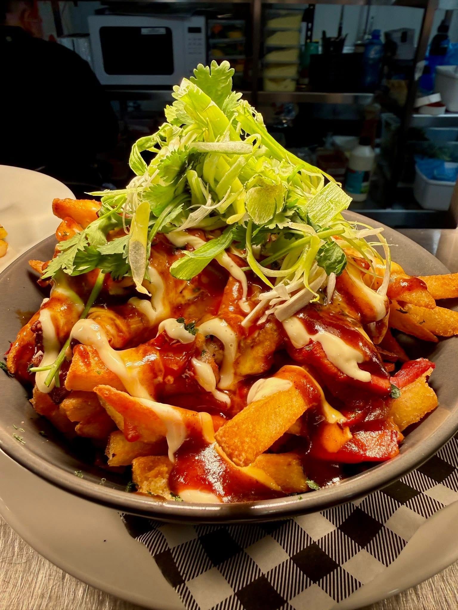 Fries topped with sauce, bacon, and celery in a gray bowl on a checkered surface.
