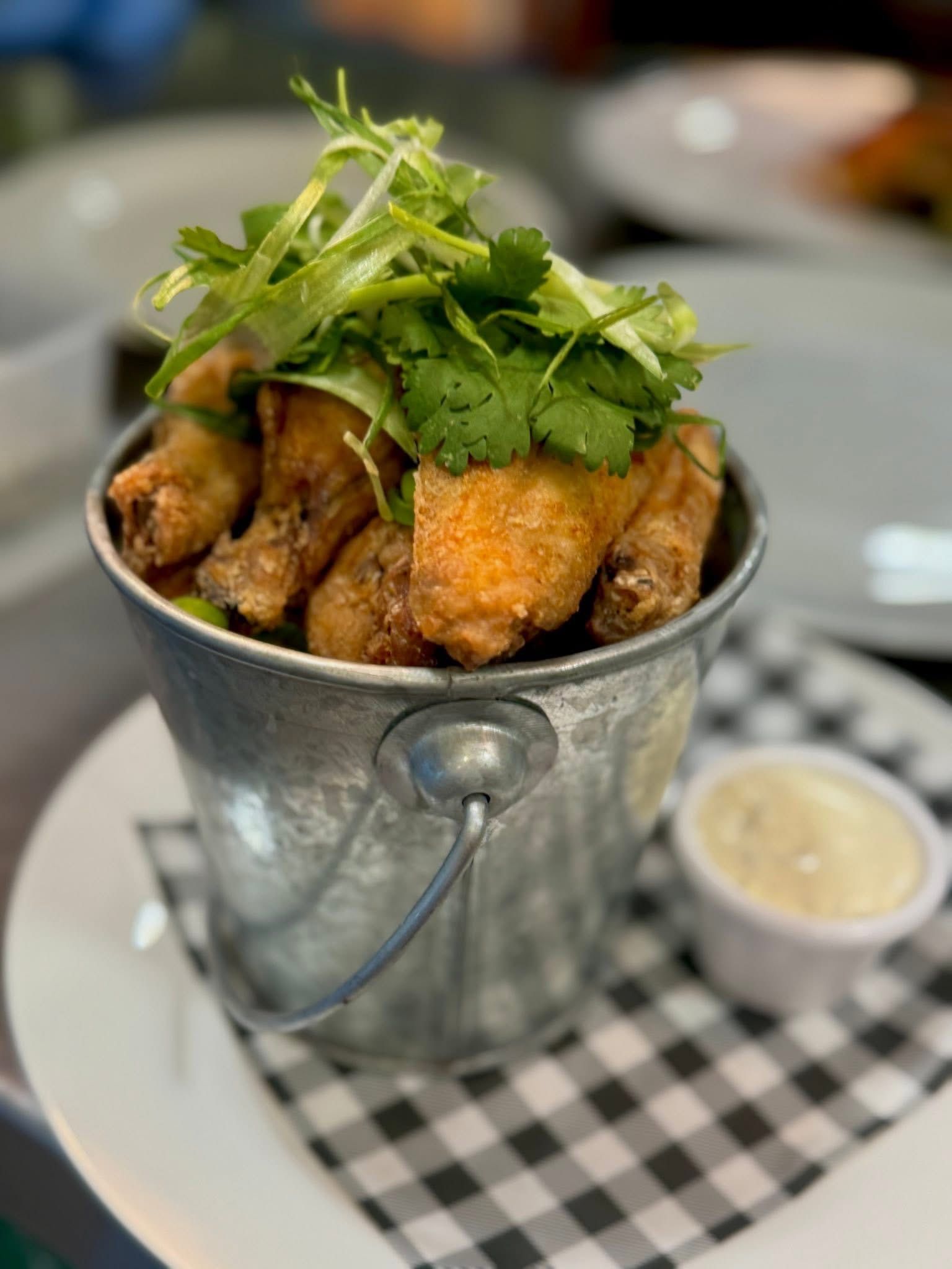 Fried chicken wings in a metal bucket, garnished with cilantro, served with sauce on a checkered napkin.