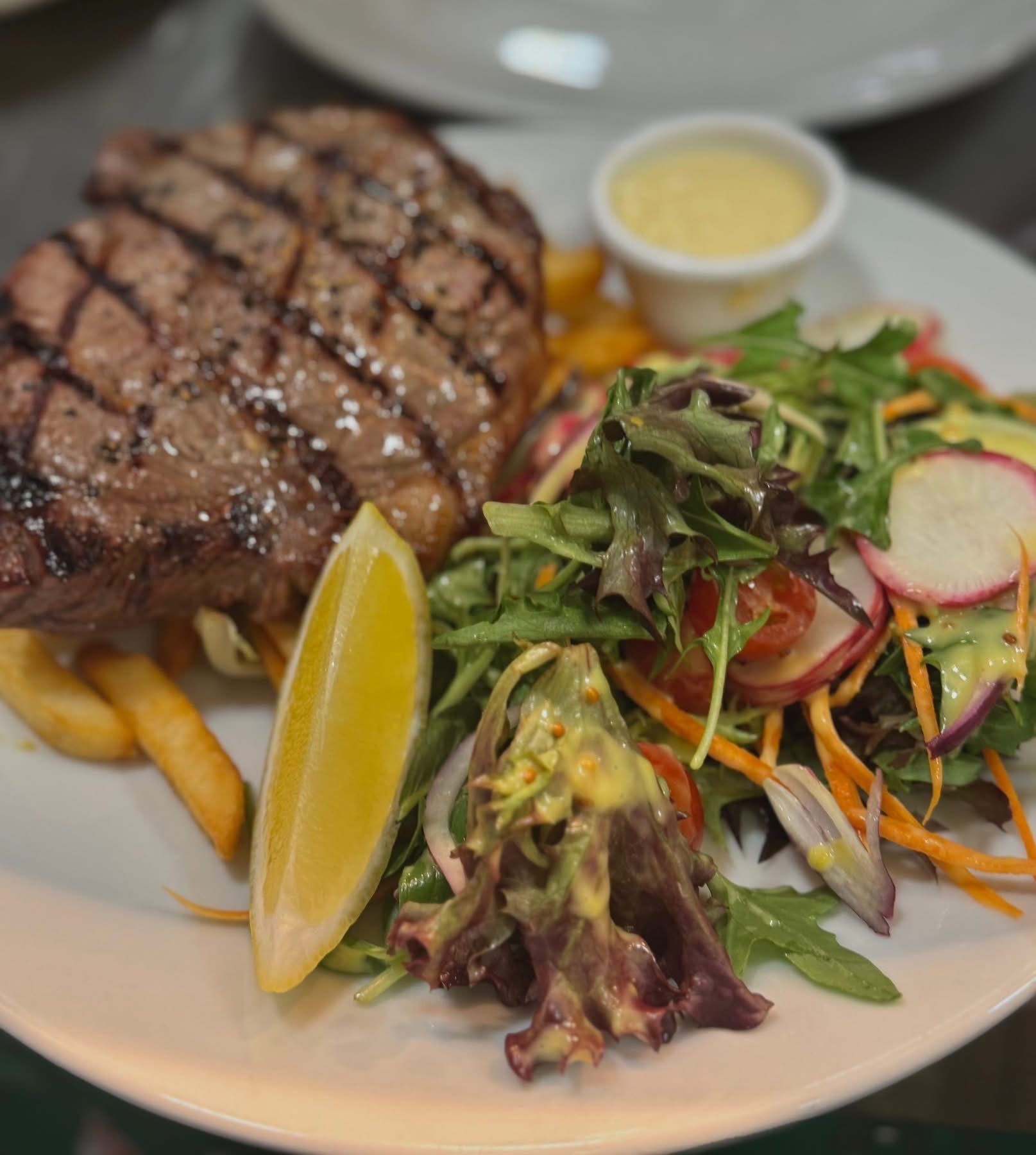 Grilled steak with fries, salad, lemon slice, and sauce on a white plate.