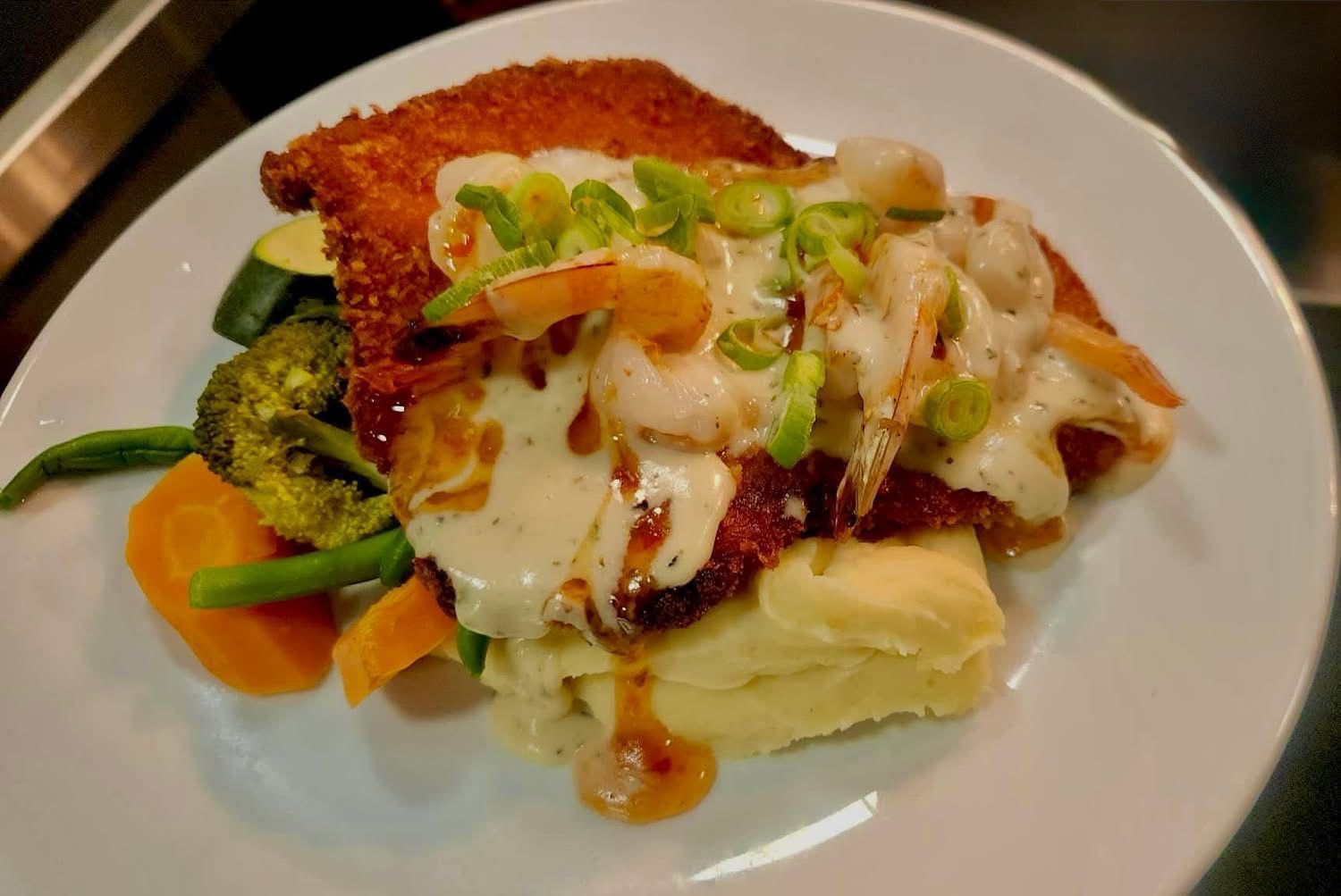Breaded entree with shrimp and sauce, over mashed potatoes, with vegetables on a plate.