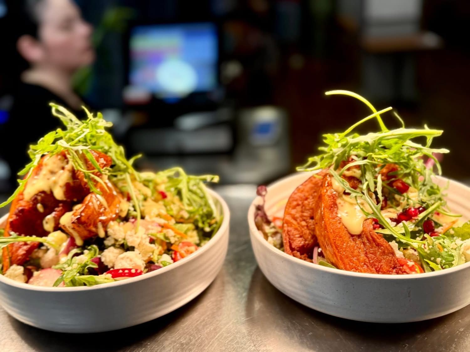 Two bowls of salad with fried food, greens, and dressing on a counter. A person in the background.