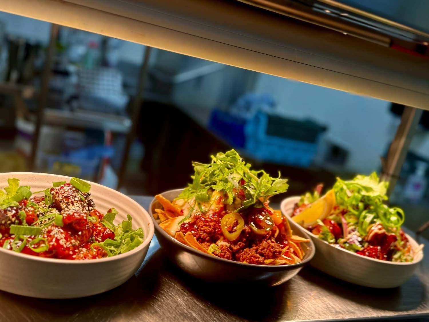 Three bowls of food, each with sauce, vegetables, and lettuce, under a warmer.