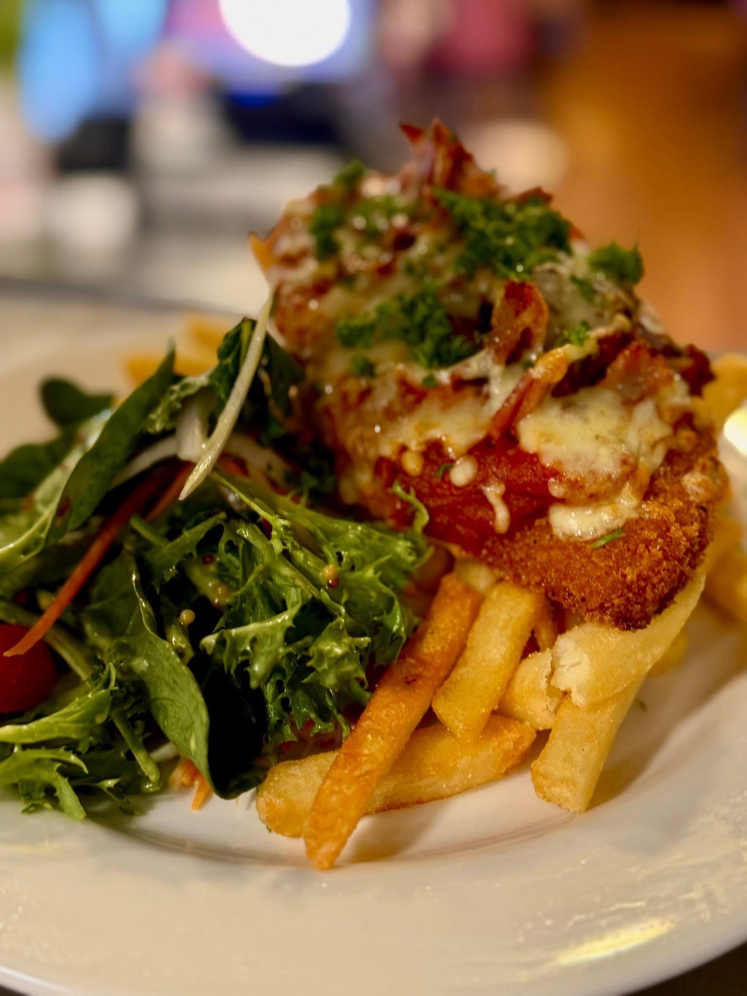 Chicken parmigiana with bacon and fries, side salad on a white plate.