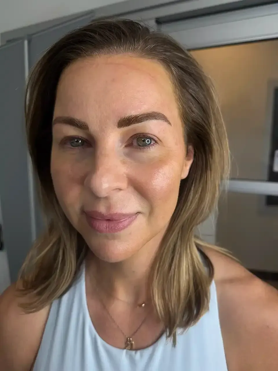 A woman with blonde hair is wearing a white tank top and a necklace.