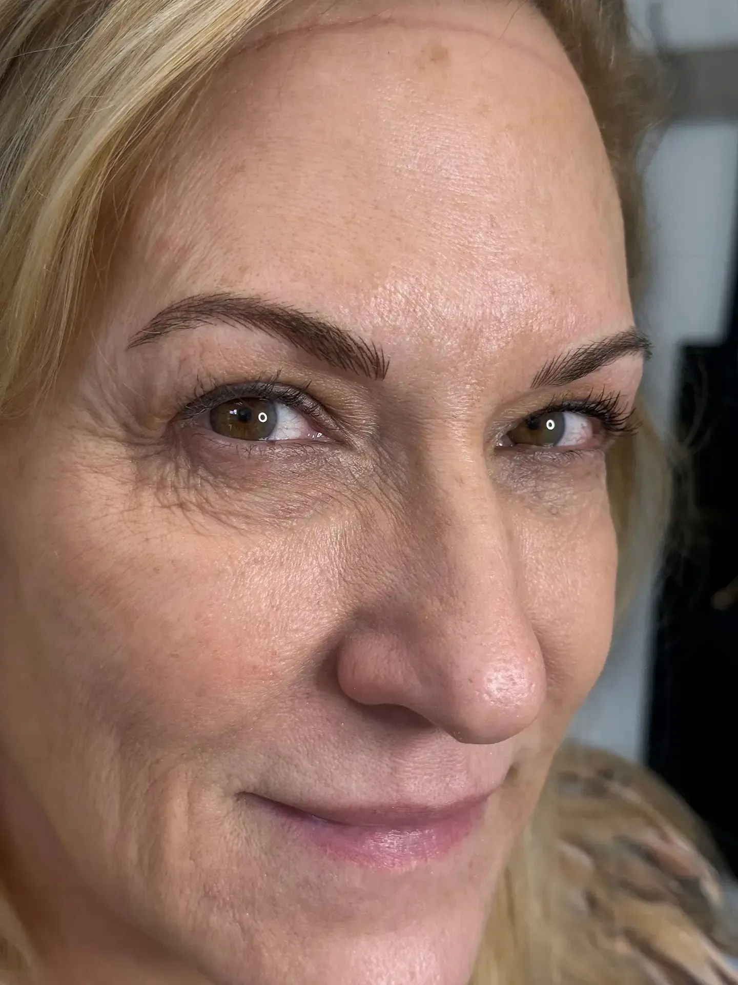 A middle-aged woman with blonde hair and brown eyebrows smiles at the camera. Her skin has visible wrinkles.