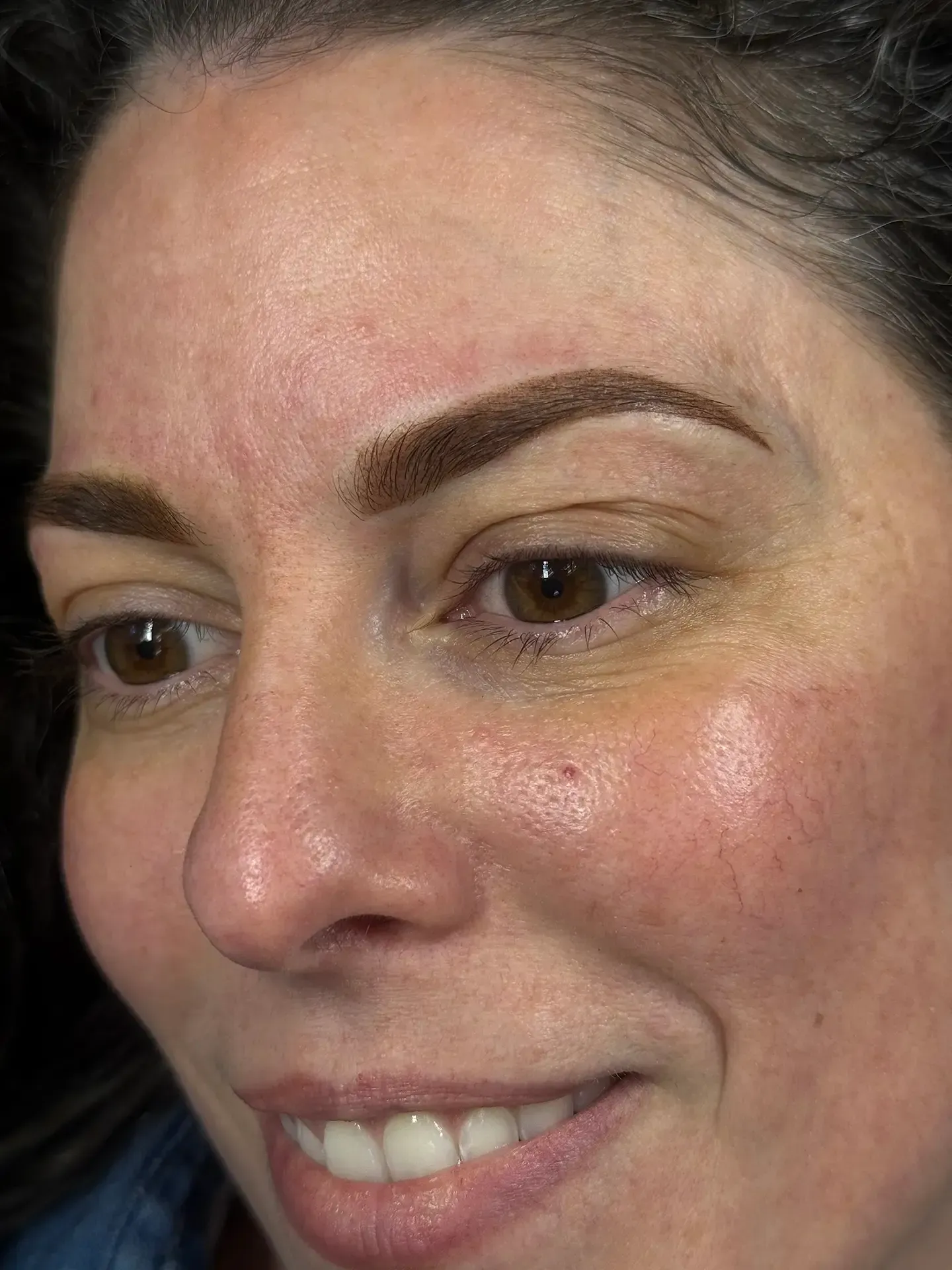 Woman with brown, filled-in eyebrows, looking down and smiling. Her skin is light with some redness, and she has brown eyes.