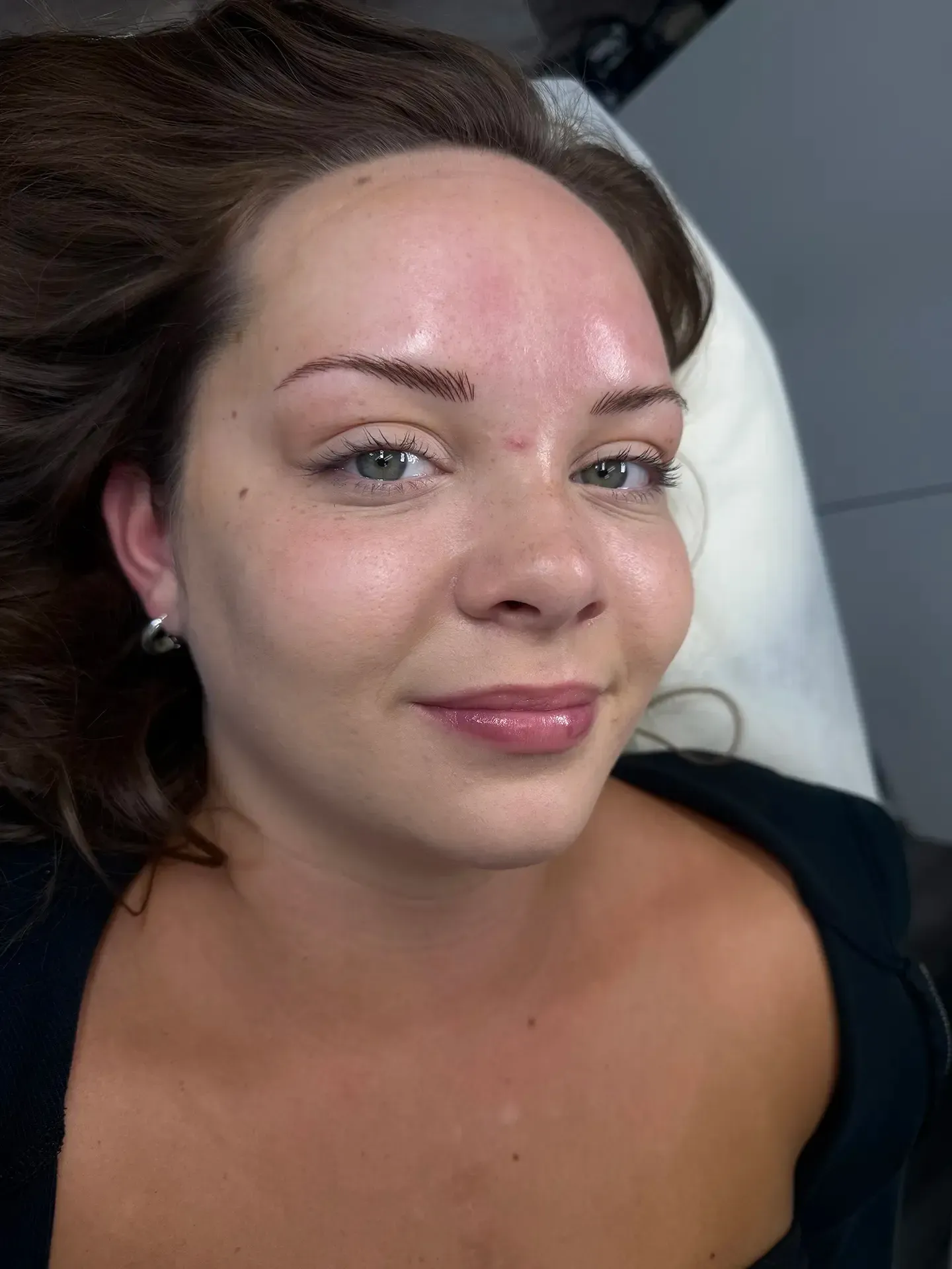 Woman with new eyebrow tattoos smiles at the camera. She is fair-skinned, with brown hair, and wearing a black top.