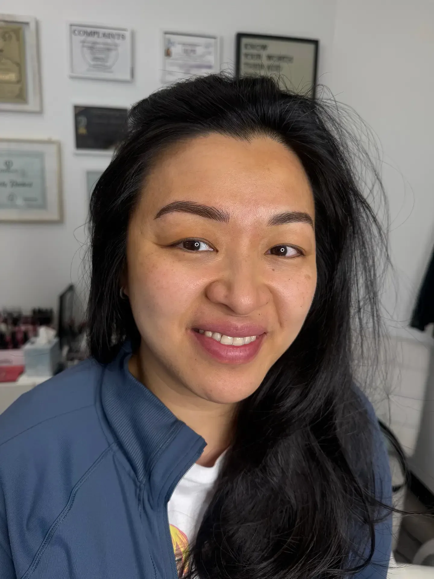 Woman with dark hair, smiling, wearing a blue jacket. Background includes framed documents, presumably in an office setting.