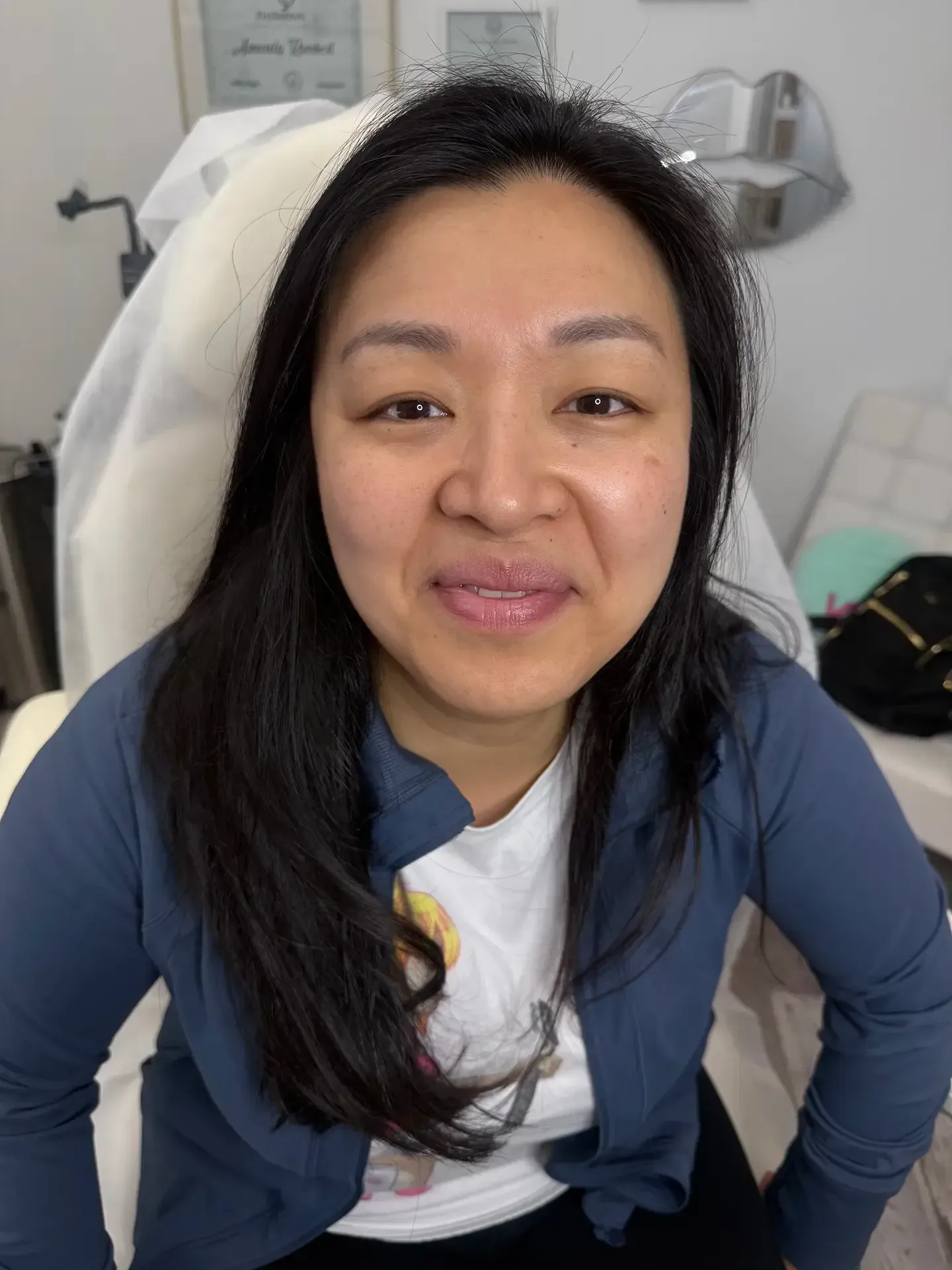 Woman with long black hair and a blue jacket smiles at the camera, sitting in a medical setting.