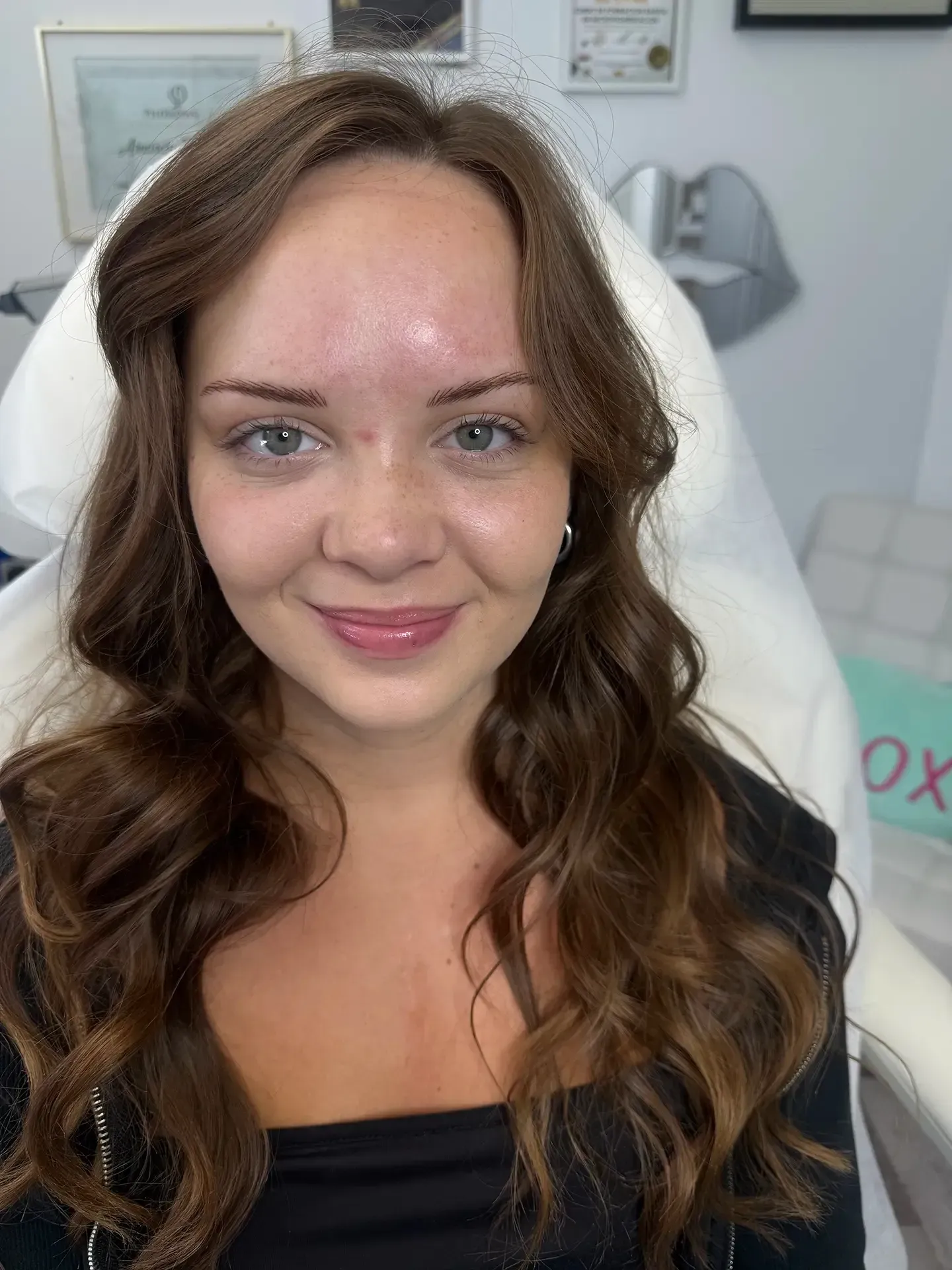 Woman with long wavy brown hair, light skin, and a slight smile, seated in what appears to be a medical or beauty office.