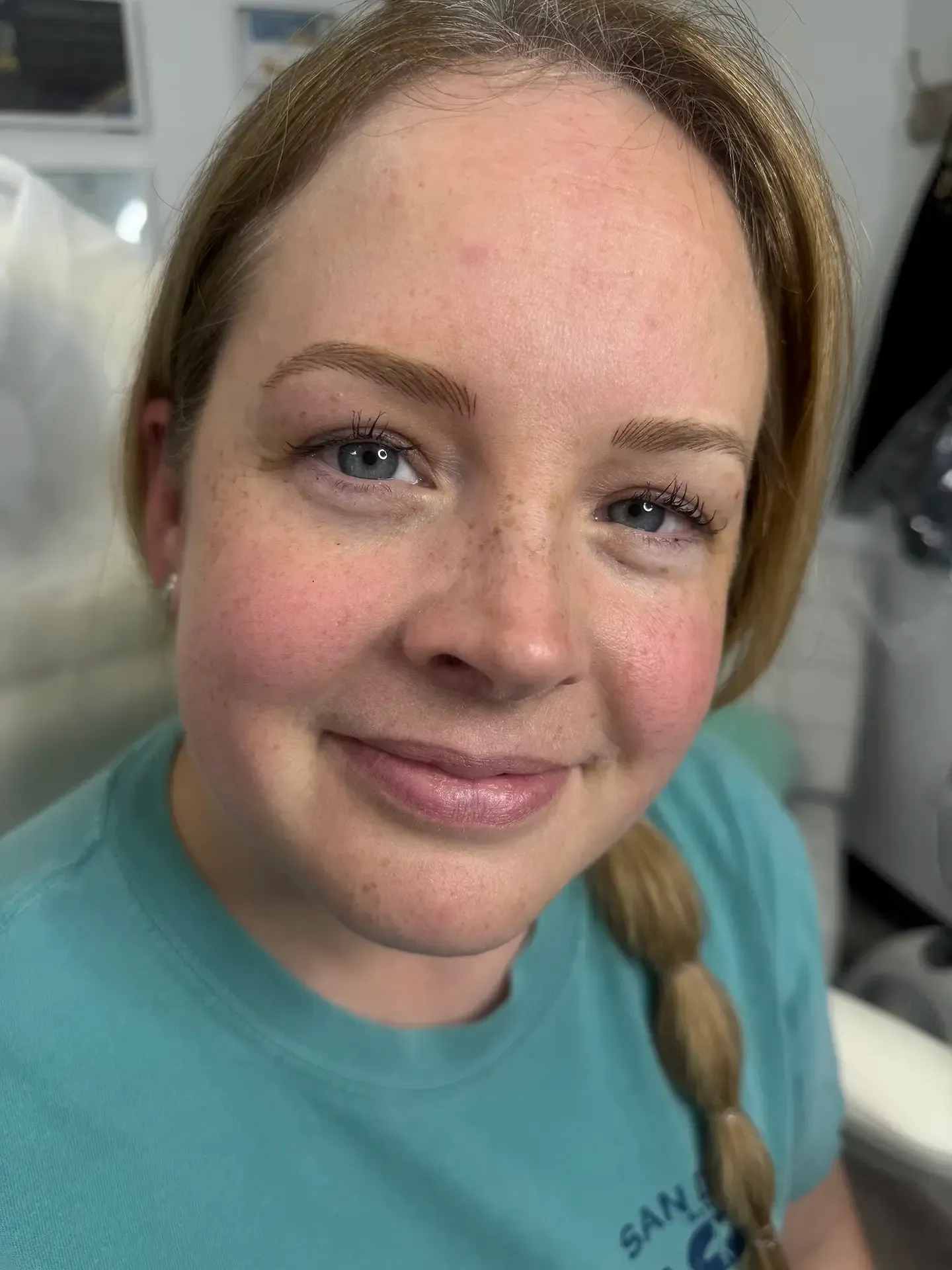 A woman with light brown eyebrows and fair skin smiles at the camera. She has a braid, rosy cheeks, and wears a teal shirt.