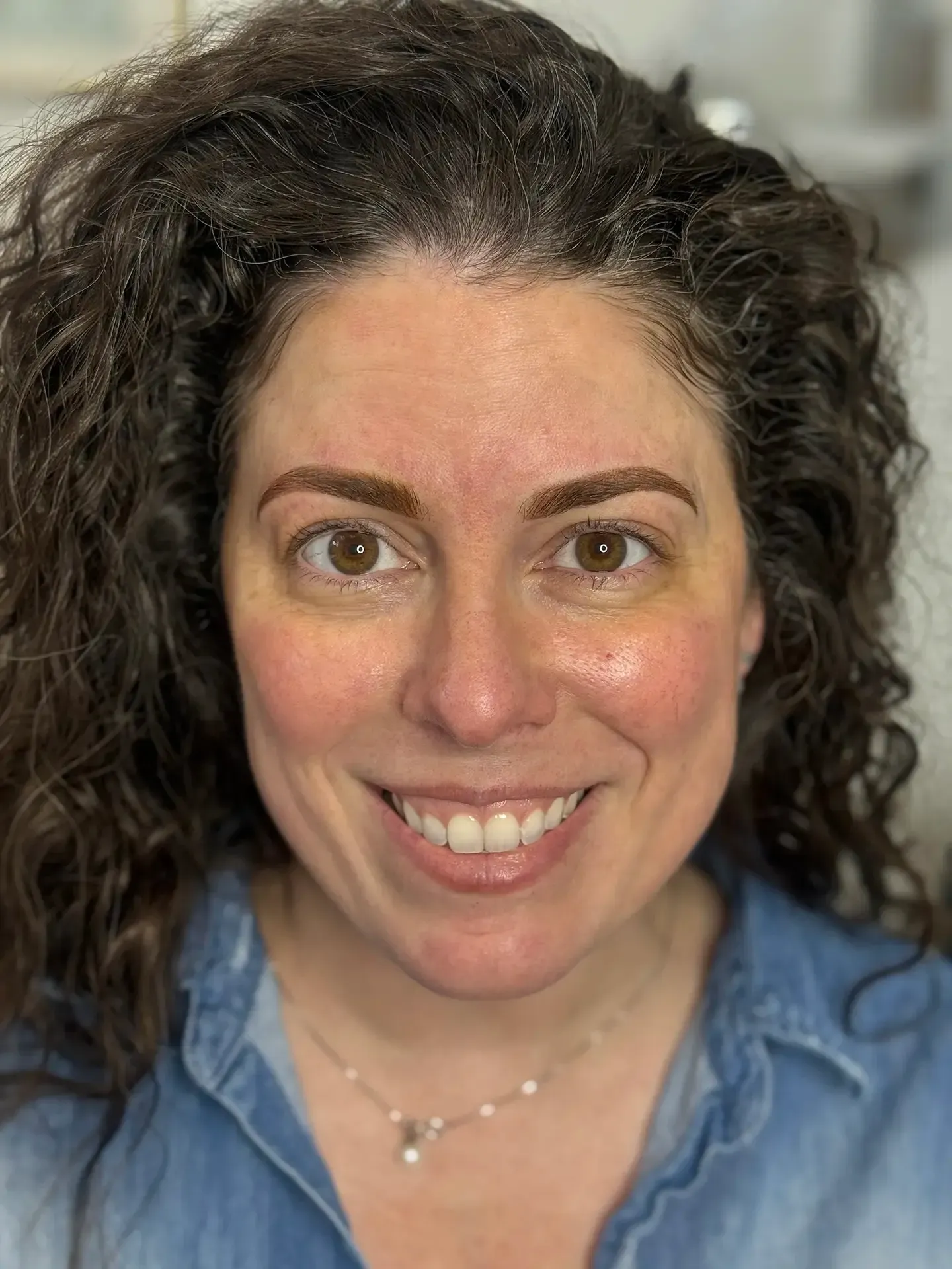 Woman with curly brown hair, smiling, wearing a denim shirt, and a pearl necklace.